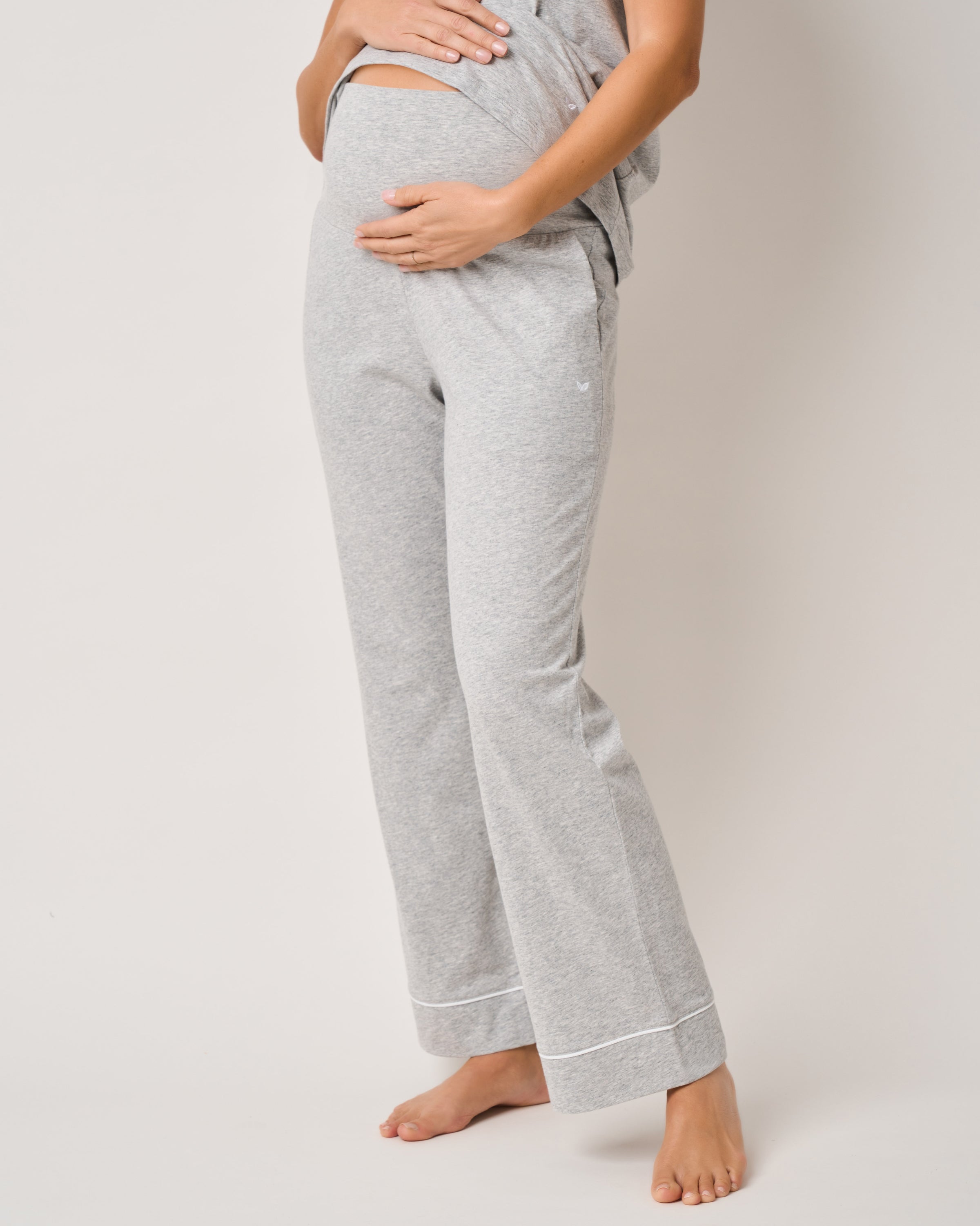 Wearing Petite Plumes Basics Maternity Set in Light Heather Grey, a person holds their belly. The set includes a loose-fitting top and wide-leg pants, projecting comfort and elegance as they stand barefoot against a plain, light backdrop.