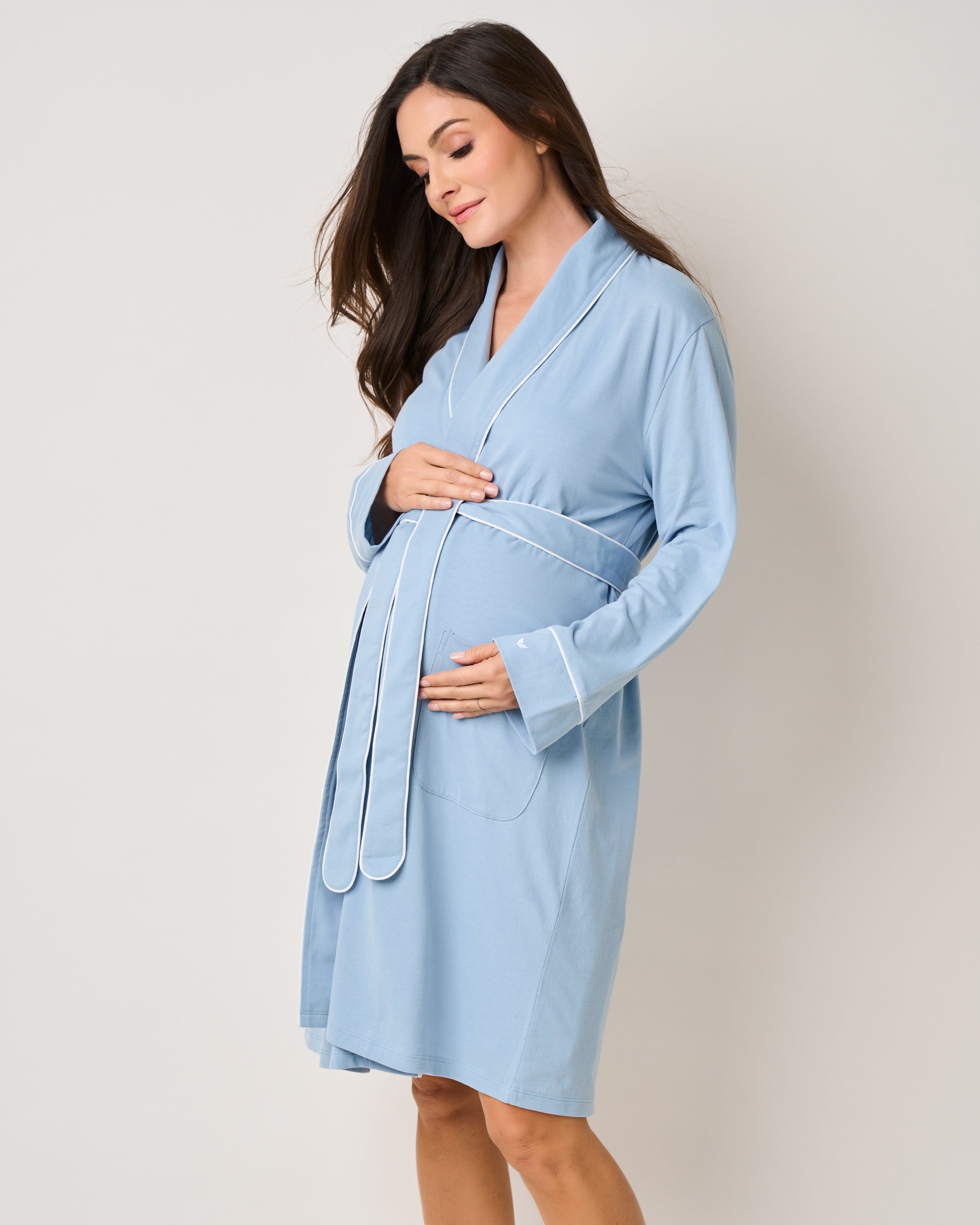 A pregnant woman stands against a plain background, gently cradling her belly in The Essential Maternity Set - Periwinkle & La Mer by Petite Plume. Her long hair flows over her shoulders as she embodies a calm and serene pregnancy journey.