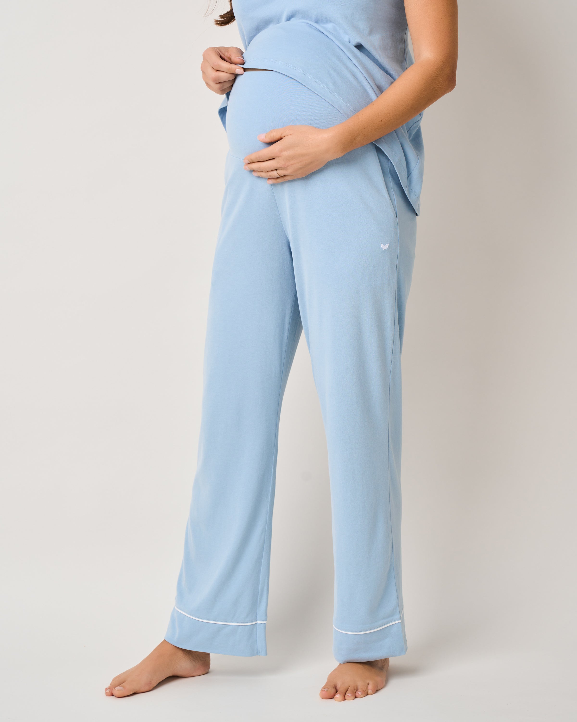 A pregnant person wears Petite Plumes The Basics Maternity Set in Periwinkle. The light blue Pima cotton pajamas showcase their baby bump, with cozy pants featuring a small logo that enhances the elegant maternity wear against a plain white background.