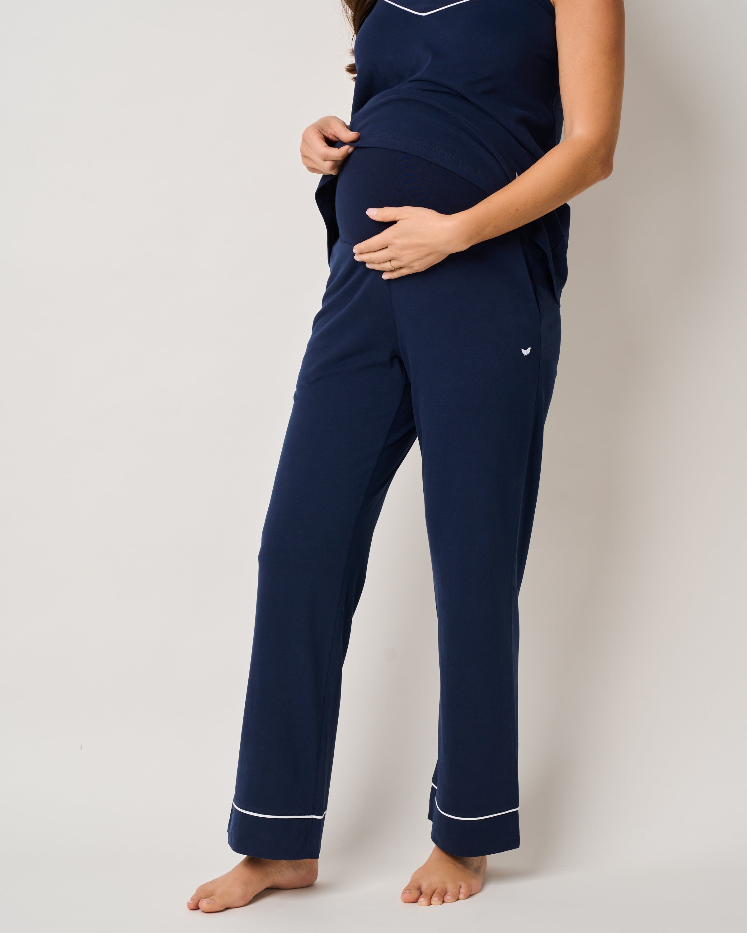 A pregnant person, barefoot on a neutral background, wears Petite Plumes Womens Pima Maternity Pants in Navy with white trim. The soft Pima cotton outfit includes a sleeveless top and full-length pants designed for comfort and support as they gently hold their belly.