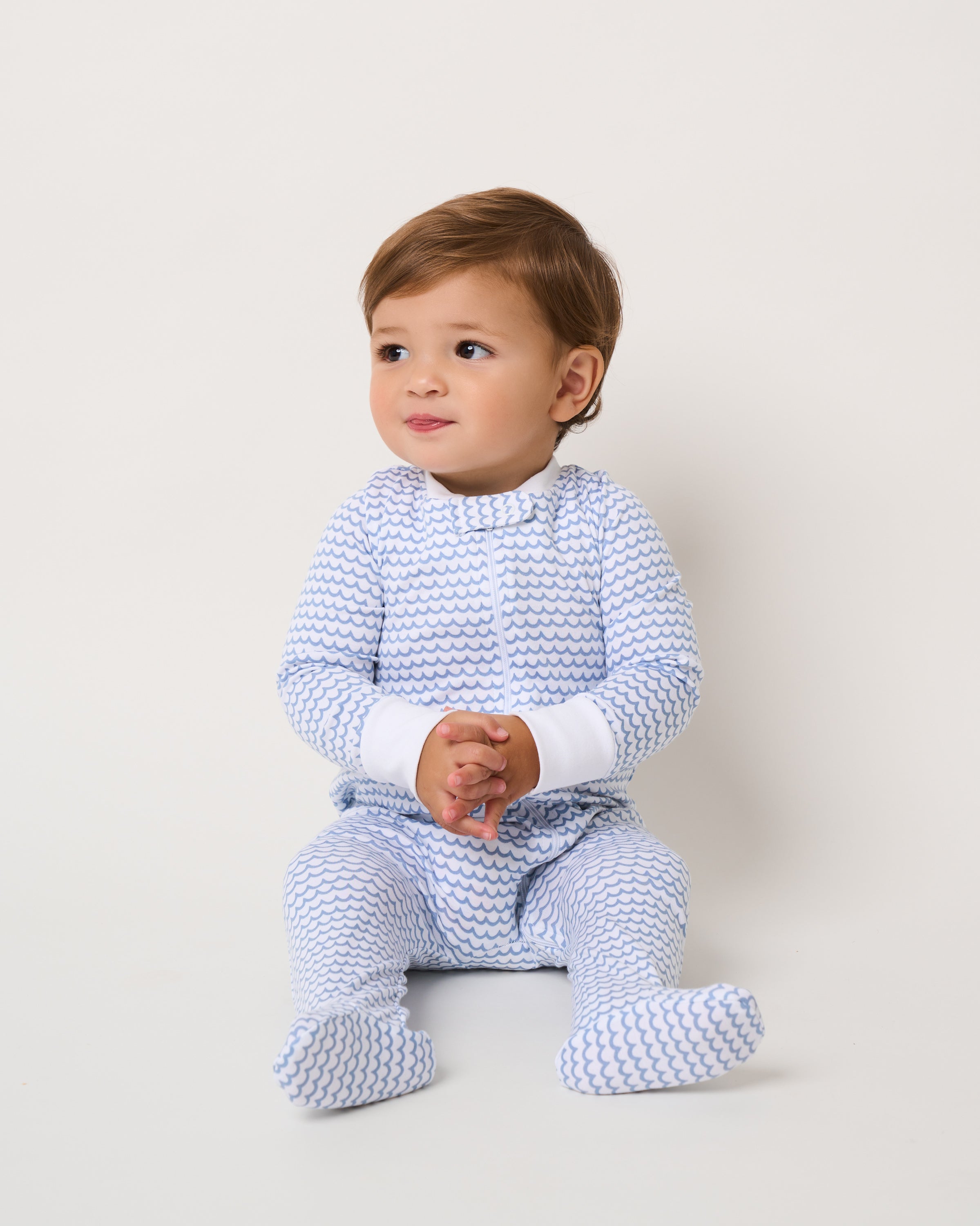 A toddler in the Babys Pima Romper in La Mer by Petite Plume, featuring vibrant colors, sits on a plain background. The child, with light brown hair, gazes curiously to the side with hands clasped together.