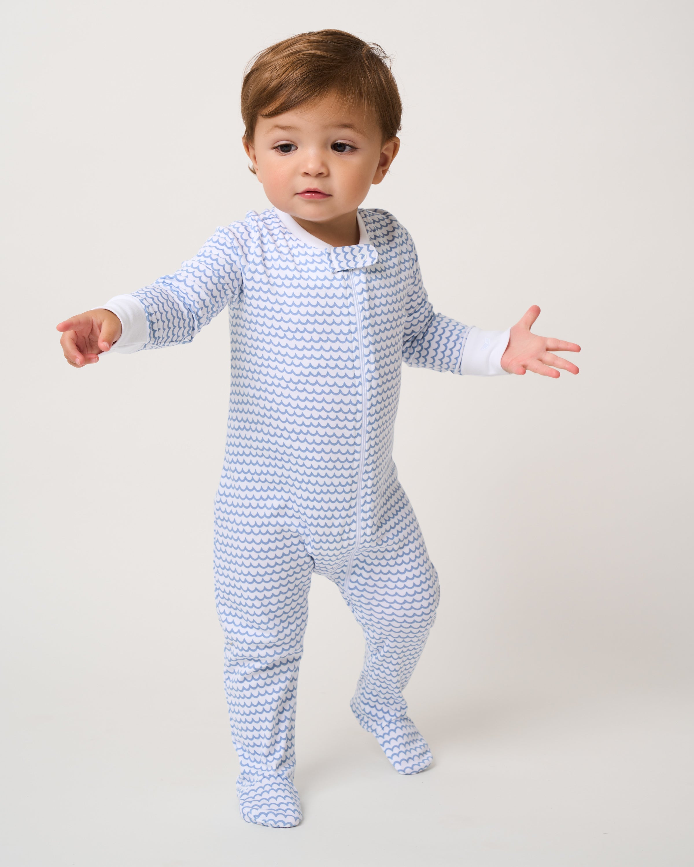 A toddler wearing the snug Babys Pima Romper in La Mer from Petite Plume stands on a plain background with slightly raised arms, sporting a curious expression as they seem to take a step forward.