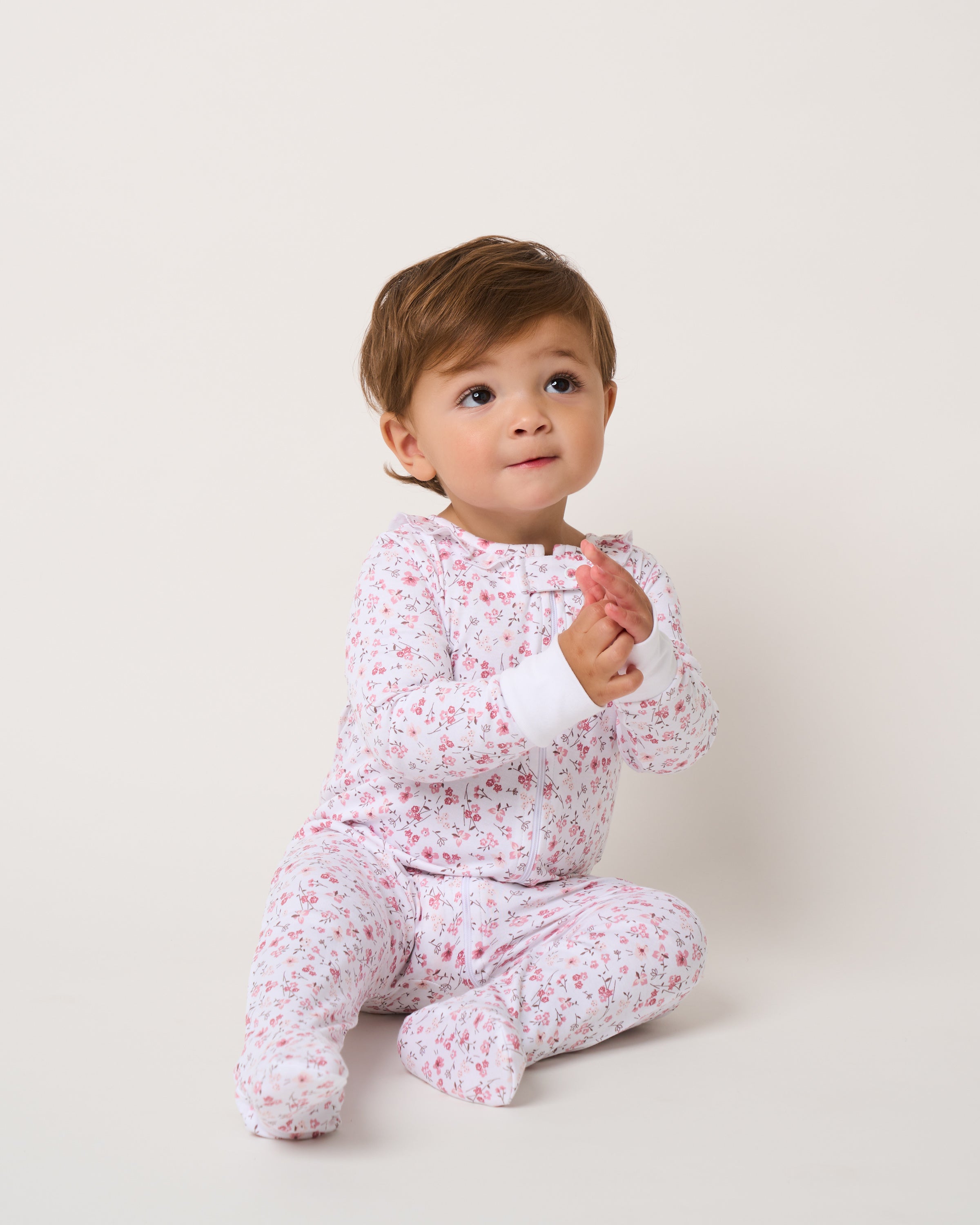 A toddler with light brown hair sits on the floor, smiling up with clasped hands while wearing Petite Plumes ultra-breathable Babys Pima Ruffle Collar Romper in Dorset Floral. The background is plain and light-colored.