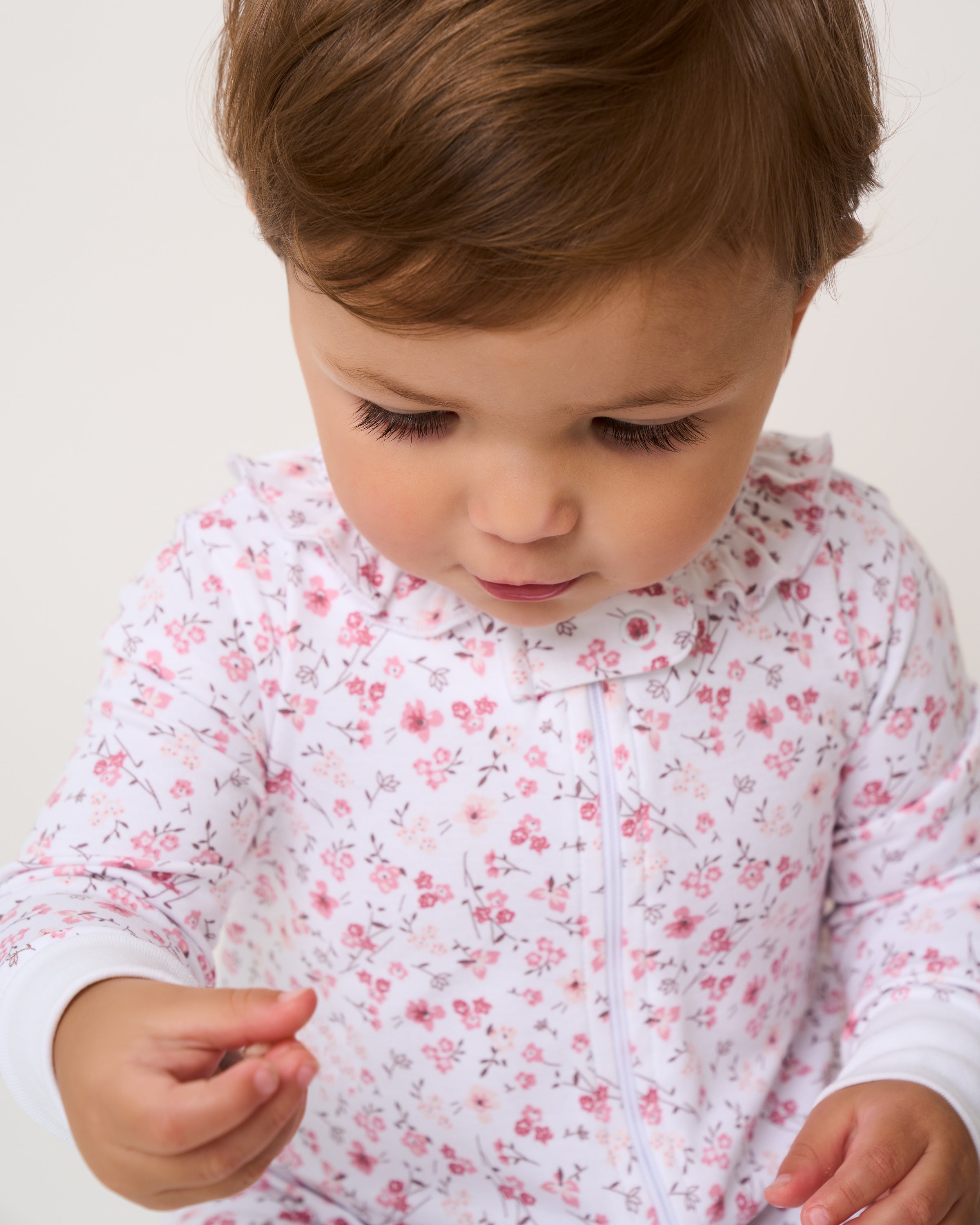 A toddler with short brown hair, wearing a luxurious Petite Plume Babys Pima Ruffle Collar Romper in Dorset Floral, looks down intently while holding a small object against a plain background.