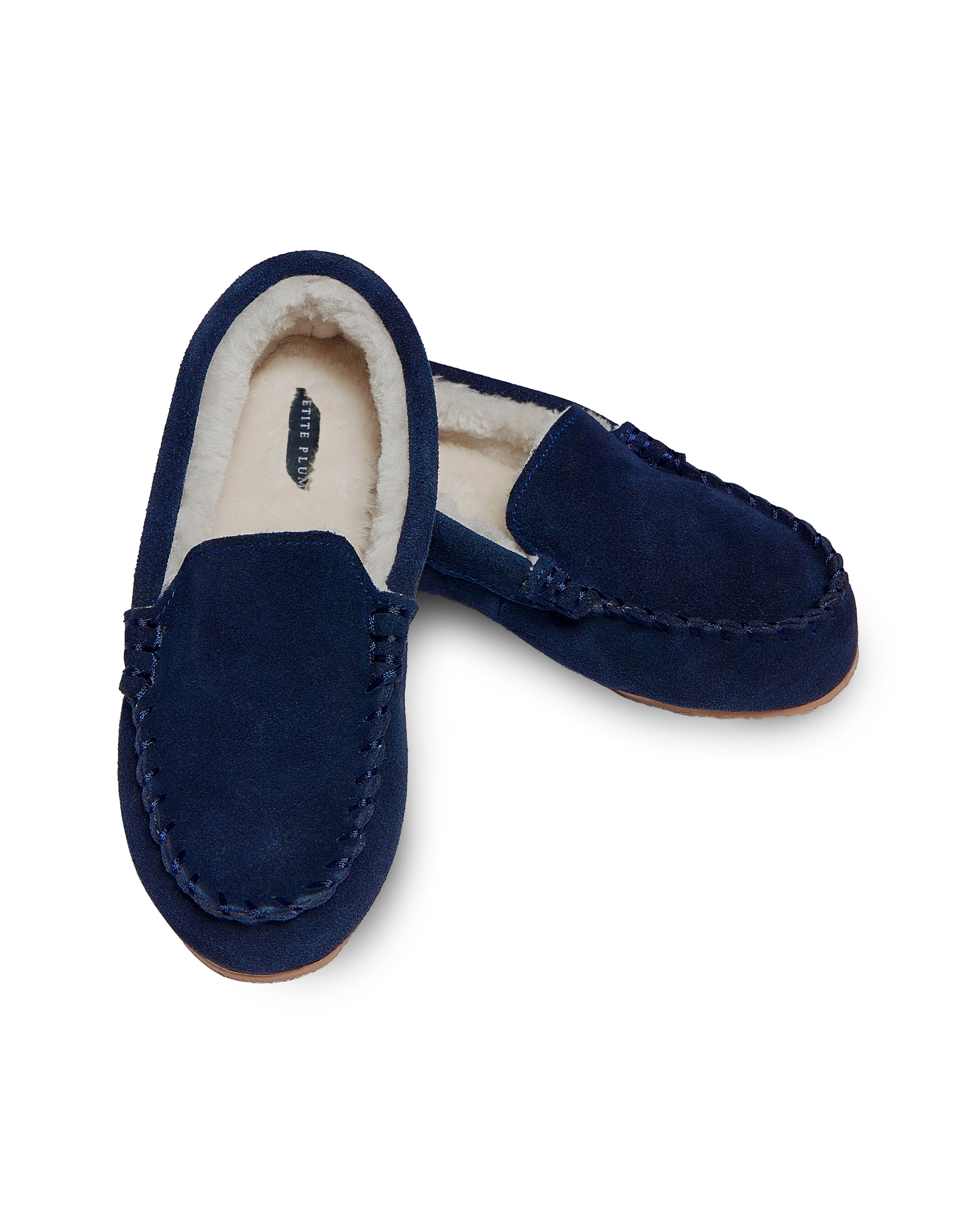 A pair of Petite Plume Kids Suede Slippers in Navy with ultra-soft faux fur lining. One slipper stands upright, and the other lies on its side, revealing the cozy interior.