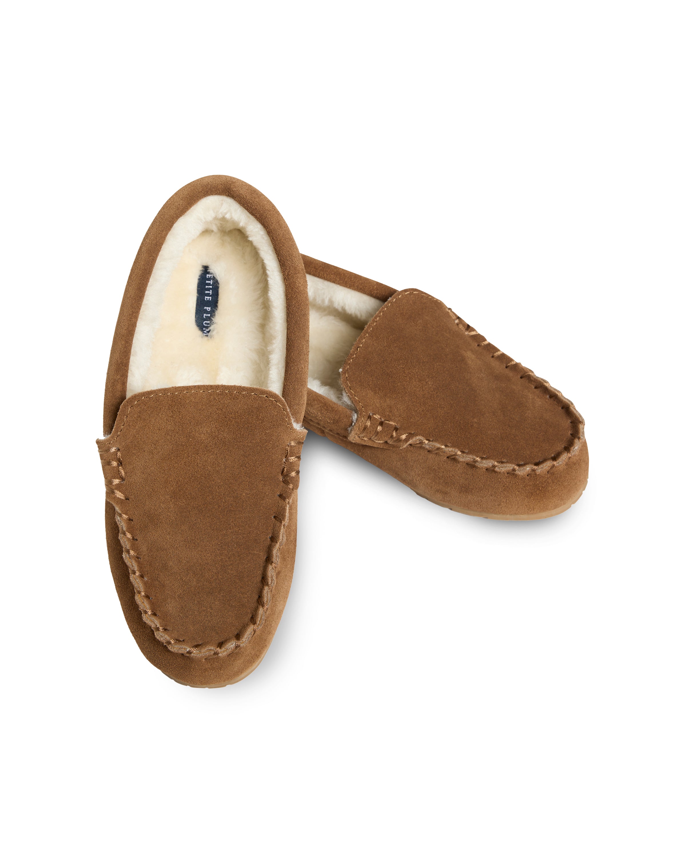 A pair of Petite Plume Kids Suede Slippers in Tan features ultra-soft suede with white faux fur lining. One slipper stands upright displaying the plush interior, while the other rests diagonally behind, inviting comfort.