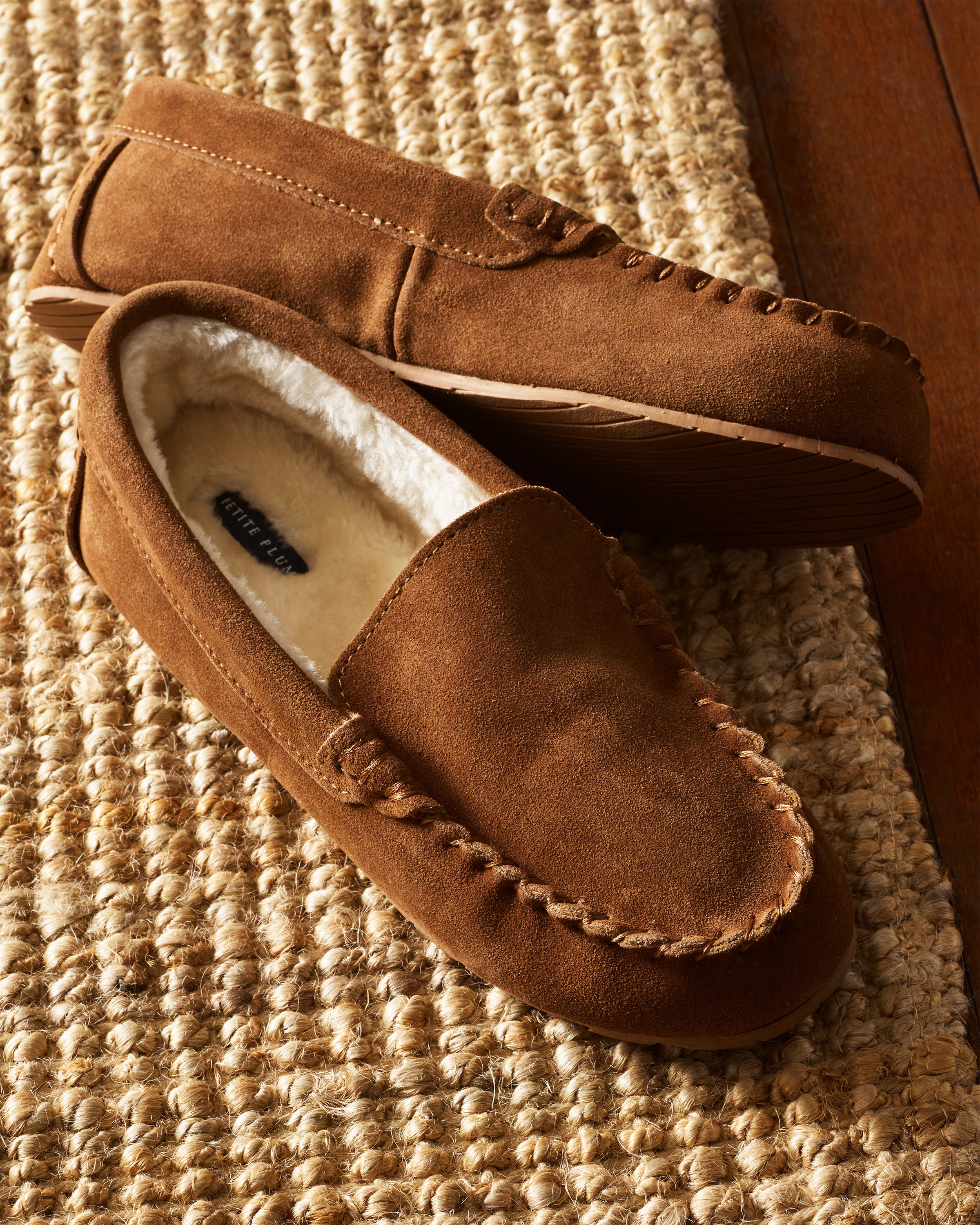 Petite Plumes Kids Suede Slippers in Tan feature a cozy ultra-soft faux fur lining and visible stitching, exuding craftsmanship while resting on a woven textured rug.