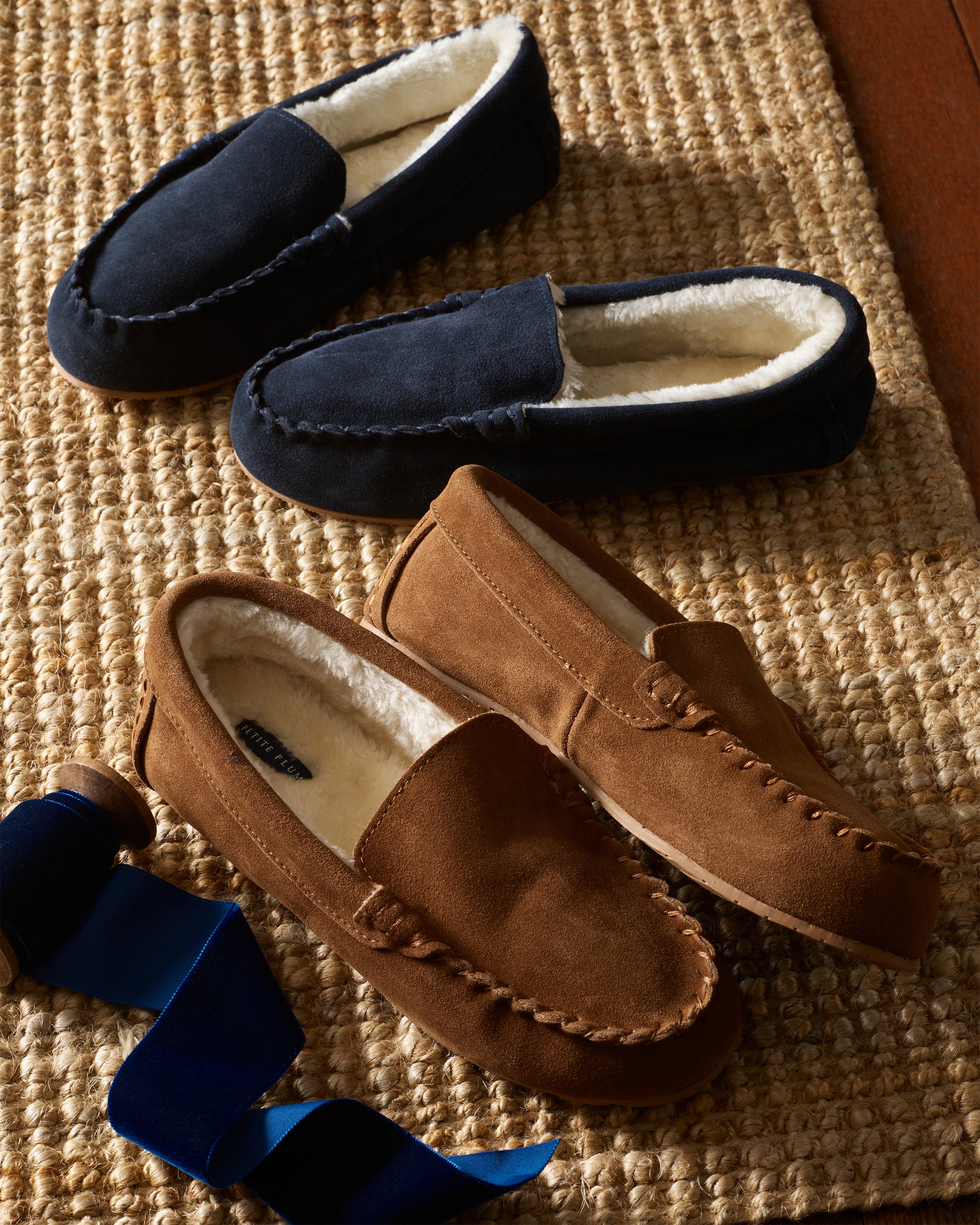 Two pairs of Petite Plume Kids Suede Slippers in tan, with ultra-soft interiors, lie on a woven rug. A spool with blue ribbon nearby adds charm to the cozy scene.