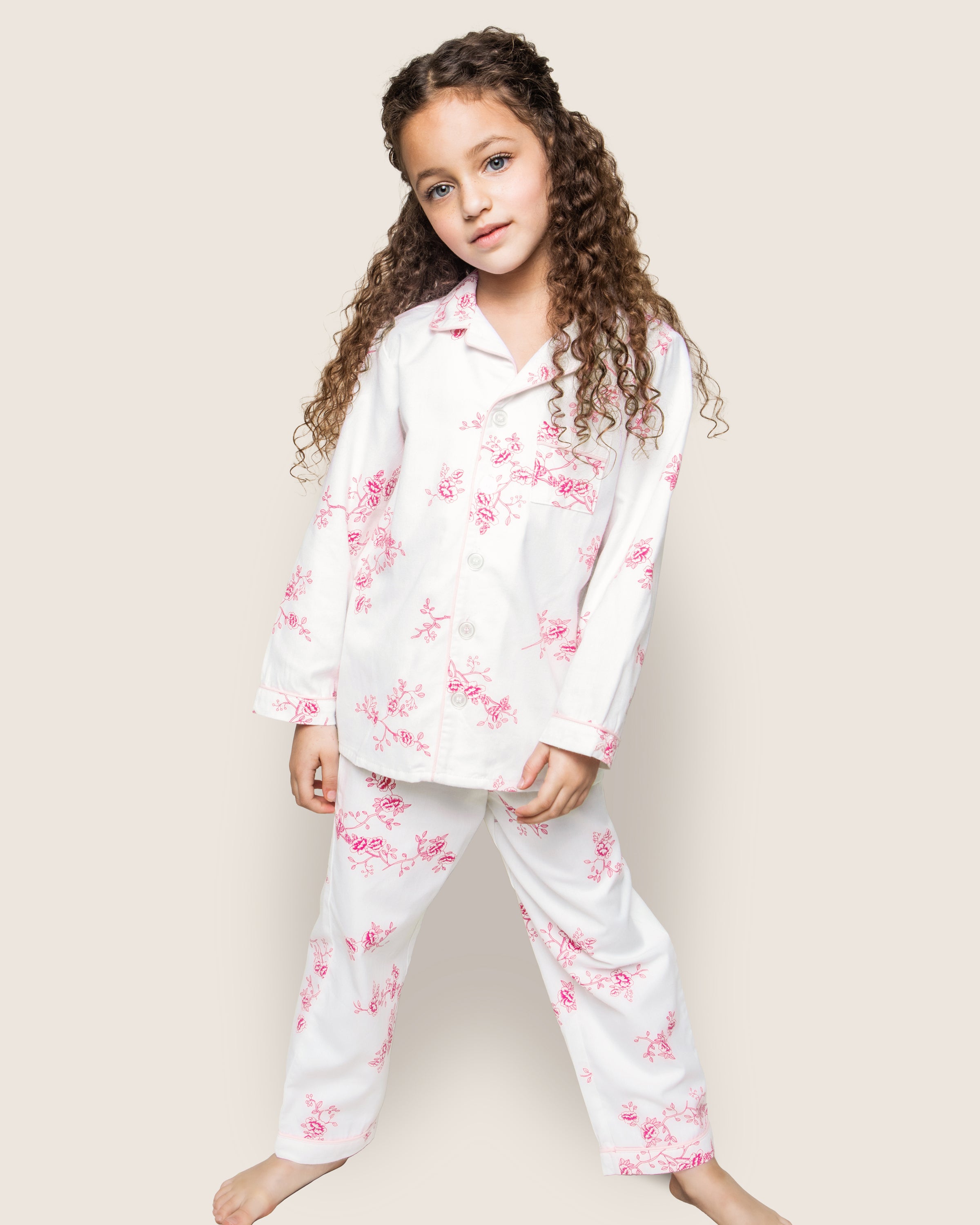 A young girl with long curly hair stands barefoot in Petite Plumes Kids Twill Pajama Set in English Rose Floral, made of luxurious cotton featuring a pink floral pattern. She poses with her head slightly tilted against a plain, light-colored background.