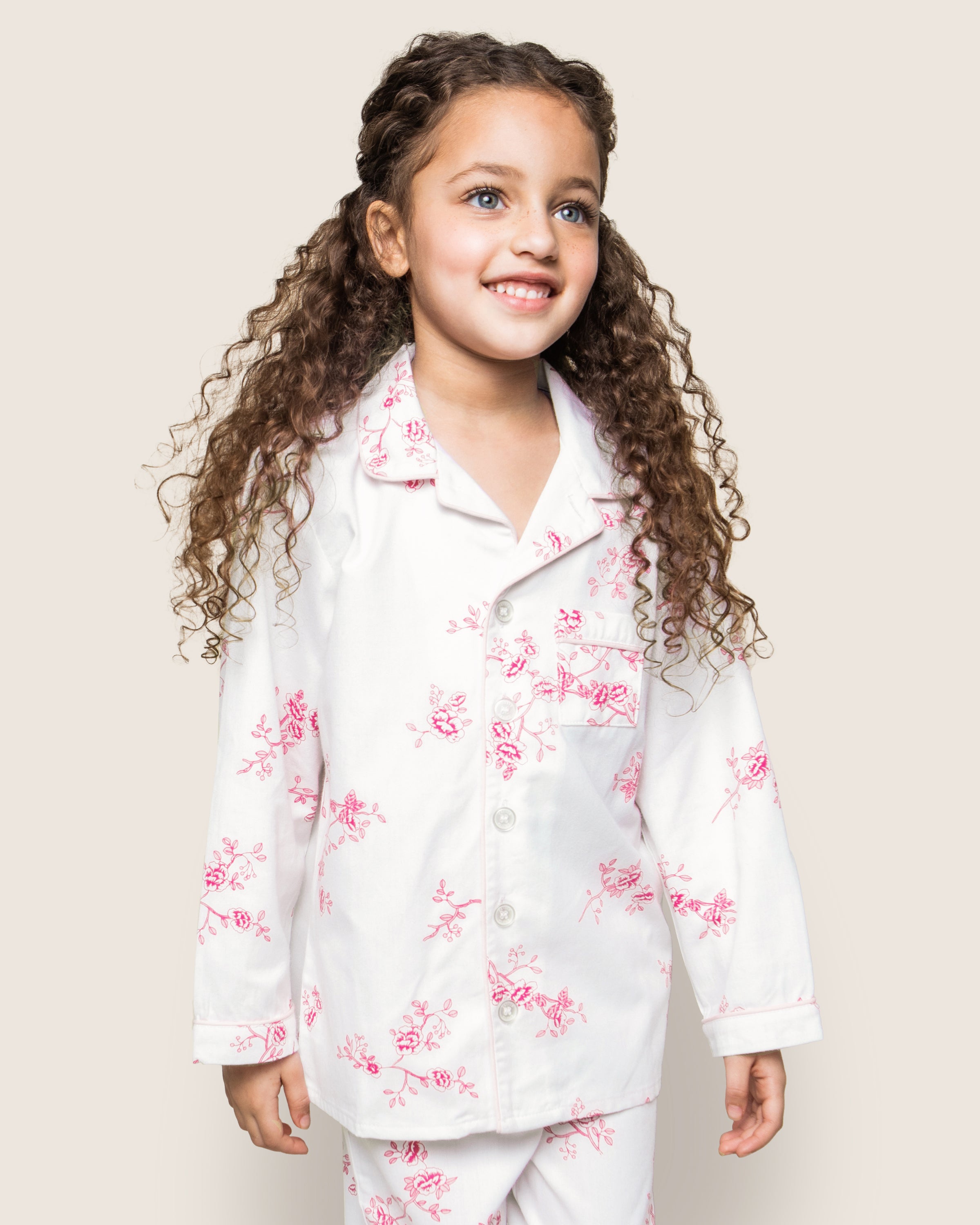 A young girl with long curly hair smiles while wearing Petite Plumes Kids Twill Pajama Set in English Rose Floral. She stands against a light beige background, exuding the charm of luxurious cotton sleepwear.