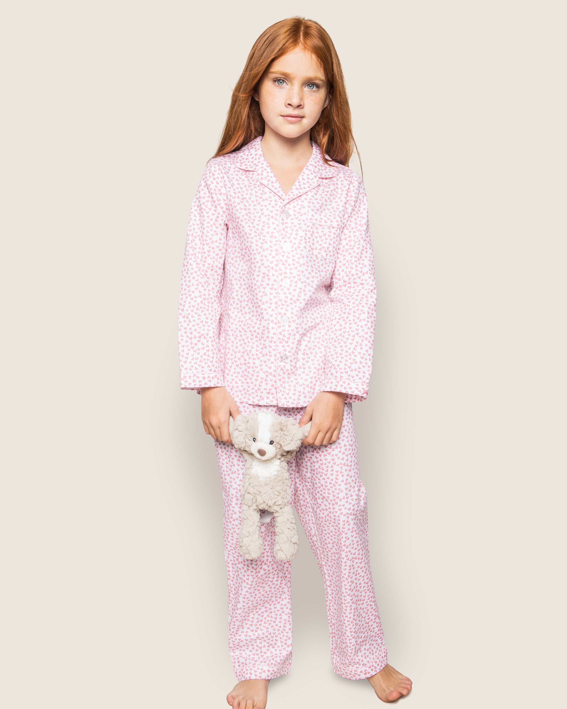 A young girl with long red hair stands against a light background, wearing Petite Plumes Kids Twill Pajama Set in Sweethearts. She holds a small white stuffed animal at her side.