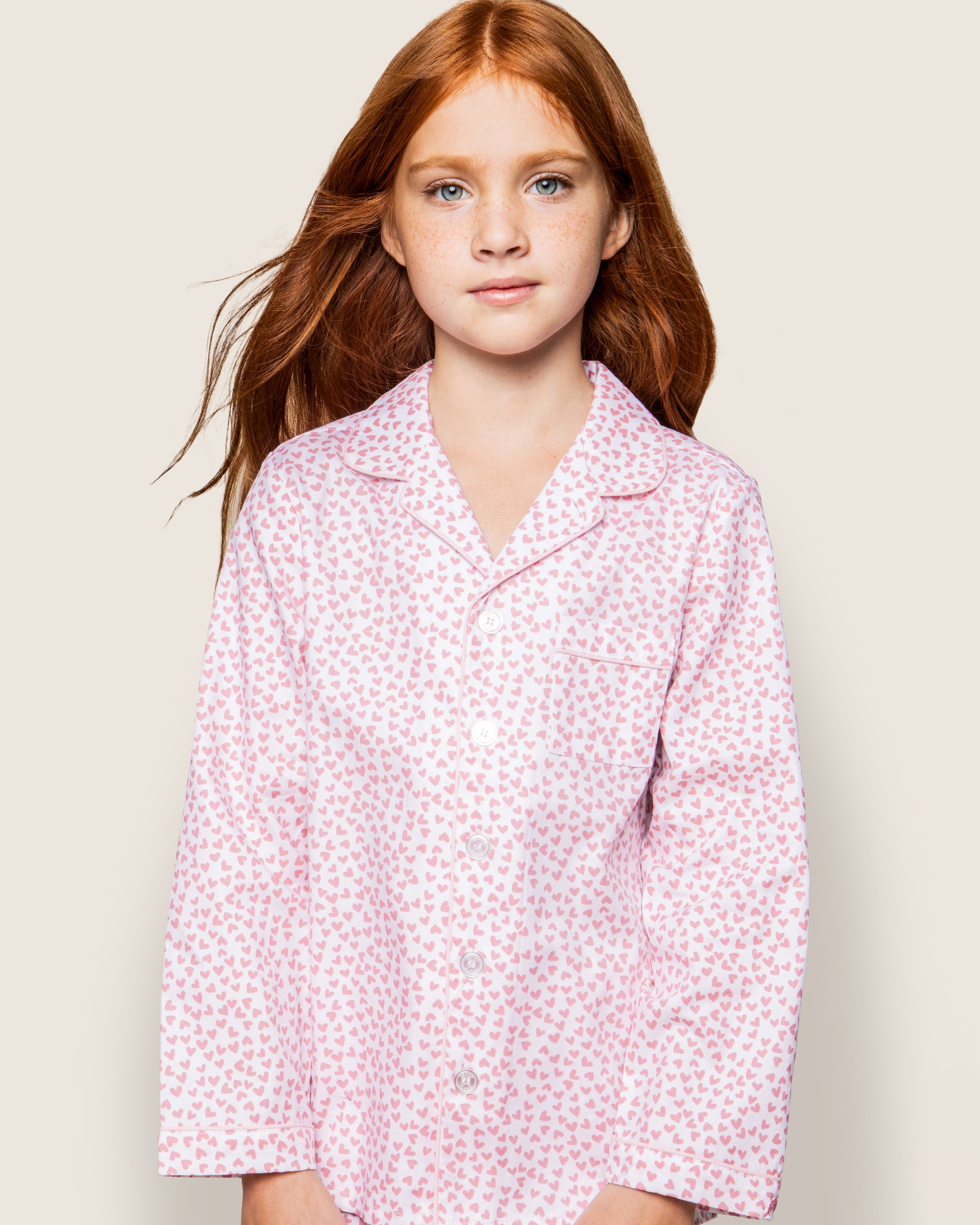 A young girl with long red hair wears a Petite Plume Kids Twill Pajama Set in Sweethearts, featuring fine cotton adorned with small red hearts. She stands against a plain, light background with a calm expression.