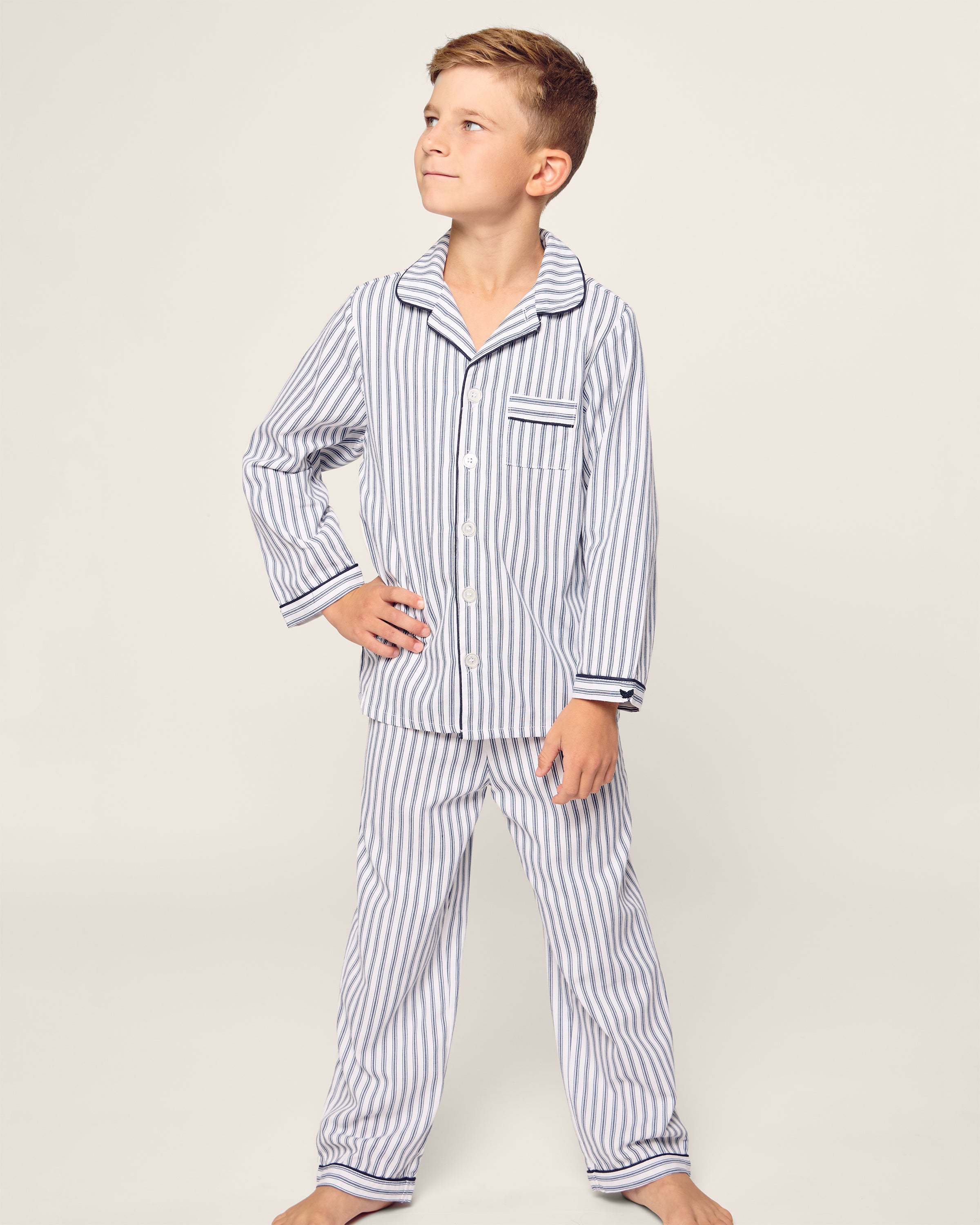 A young boy stands confidently with hands on hips in a Petite Plume Kids Twill Pajama Set in Navy French Ticking. Made from yarn-dyed cotton, the set includes a collar, front buttons, and a chest pocket. Hes barefoot and gazes to the side against a simple, light background.