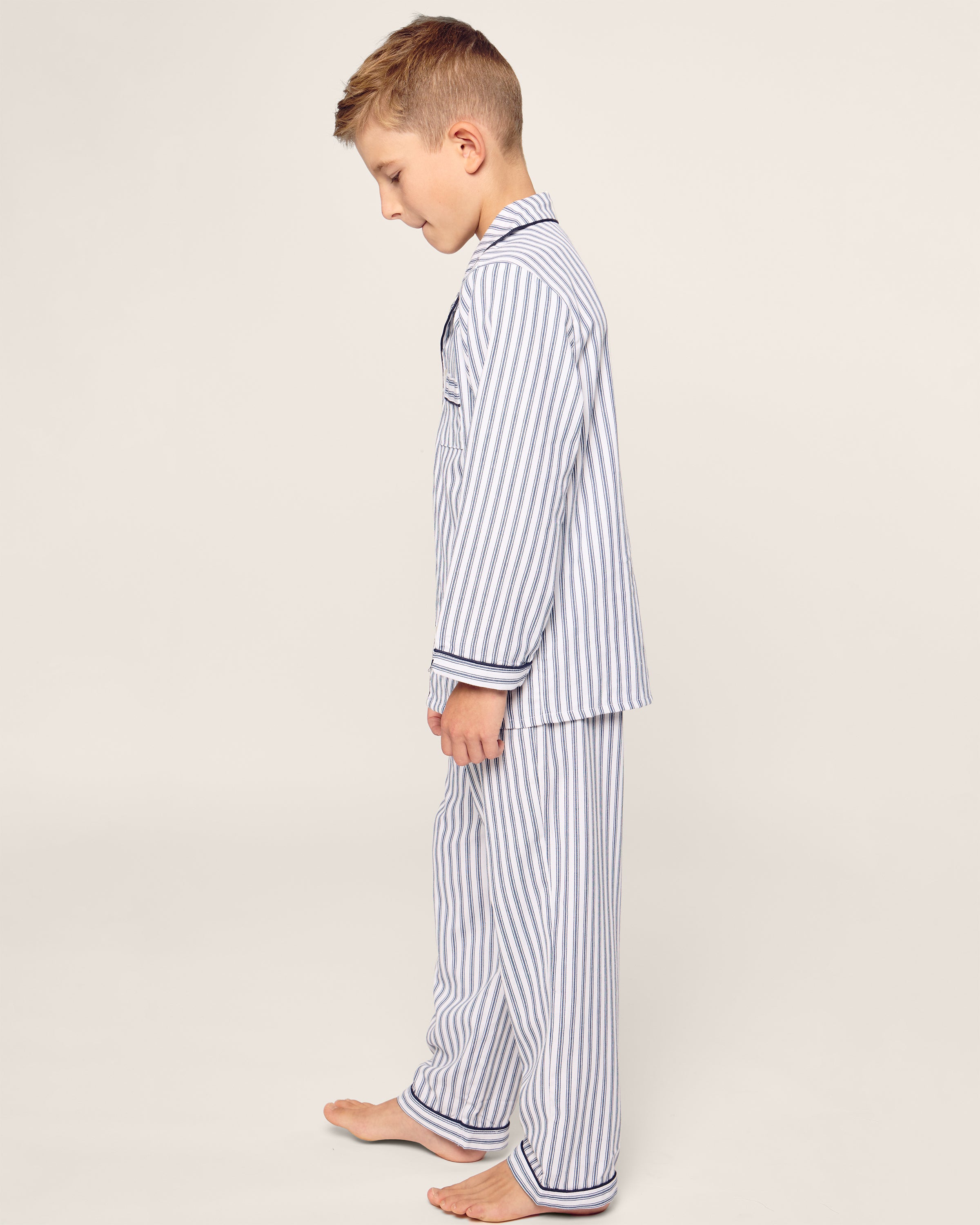 A young boy stands barefoot in a classic Petite Plume Kids Twill Pajama Set in Navy French Ticking, set against a plain background.