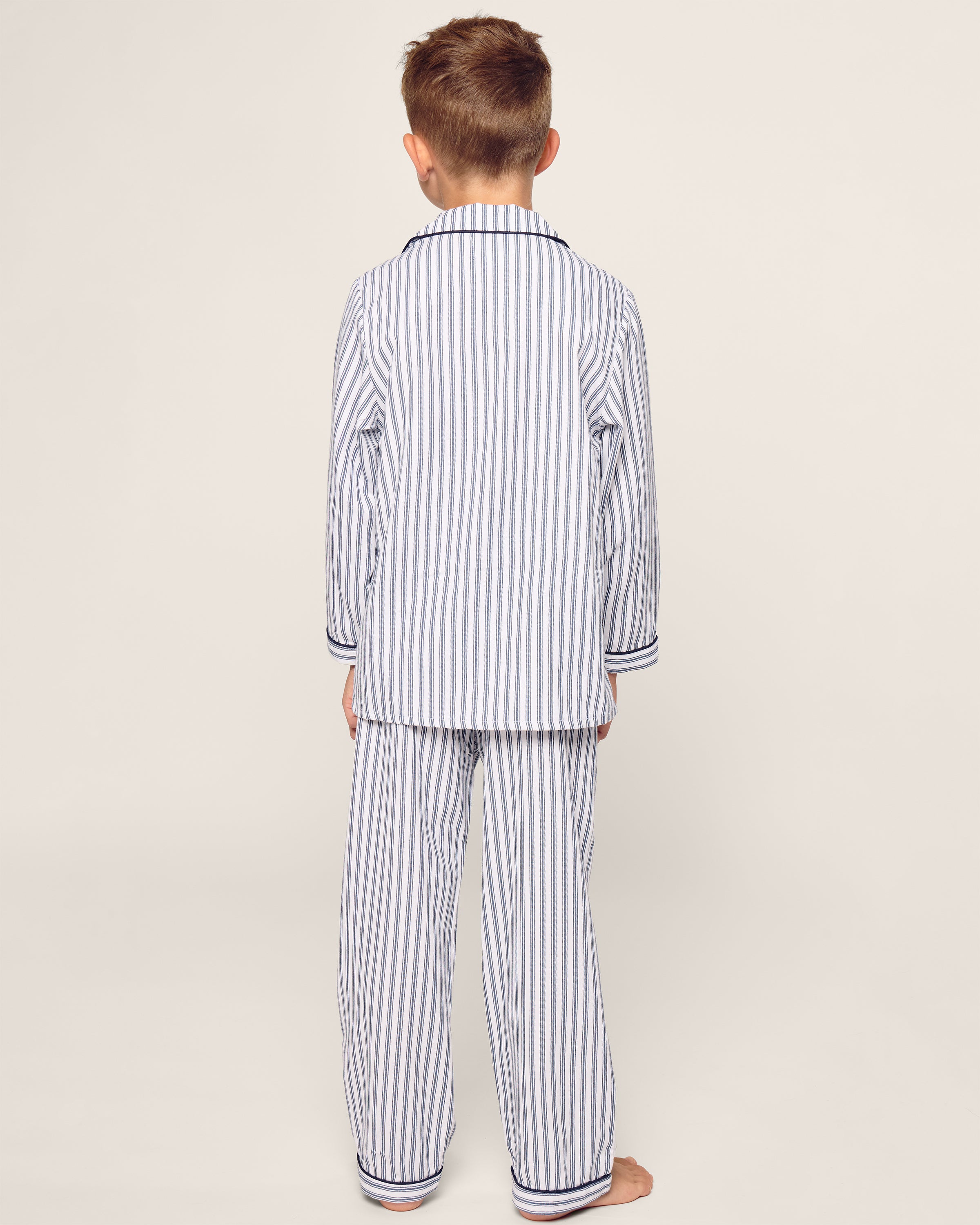 A young boy stands with his back to the camera, wearing Petite Plumes Kids Twill Pajama Set in Navy French Ticking, featuring luxurious yarn-dyed cotton fabric against a plain light beige background.