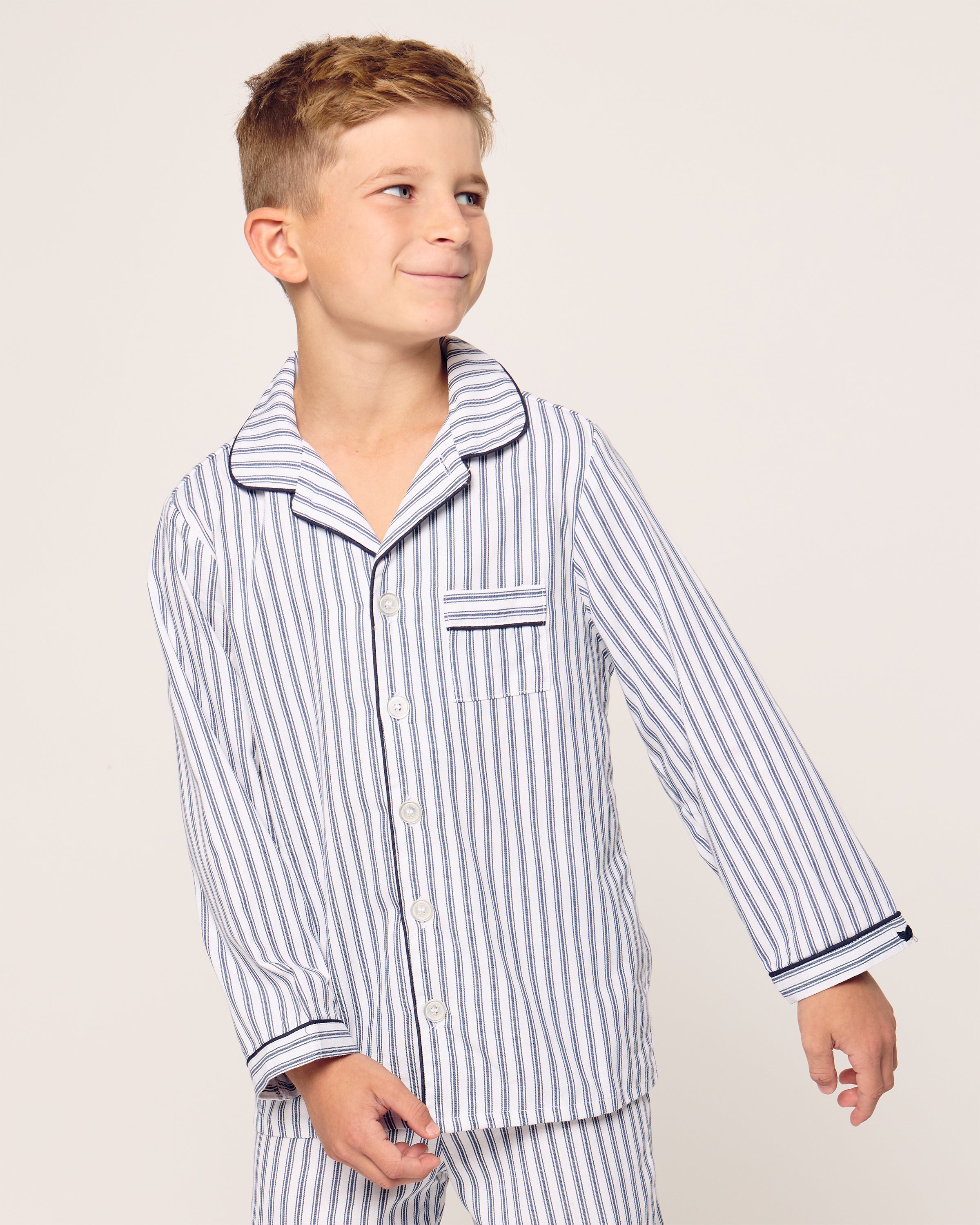 A young boy in a Petite Plume Kids Twill Pajama Set, featuring yarn-dyed navy French ticking stripes, stands against a plain background and looks to his right with a slight smile.