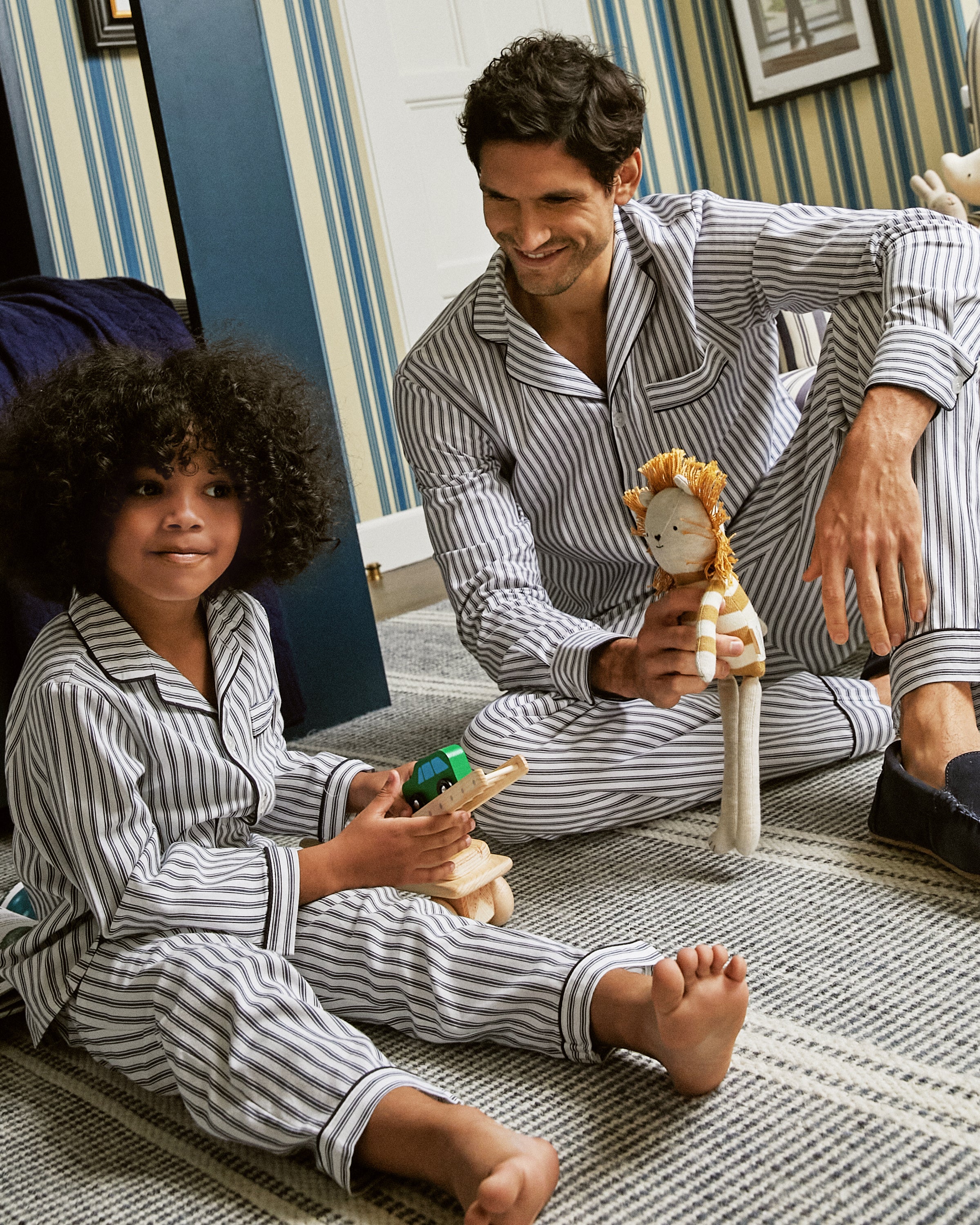 A child in Petite Plumes Kids Twill Pajama Set in Navy French Ticking plays with blocks on the carpet, while an adult also in French ticking pajamas holds a stuffed lion. The blue wallpaper with white stripes highlights their cozy outfits beautifully.