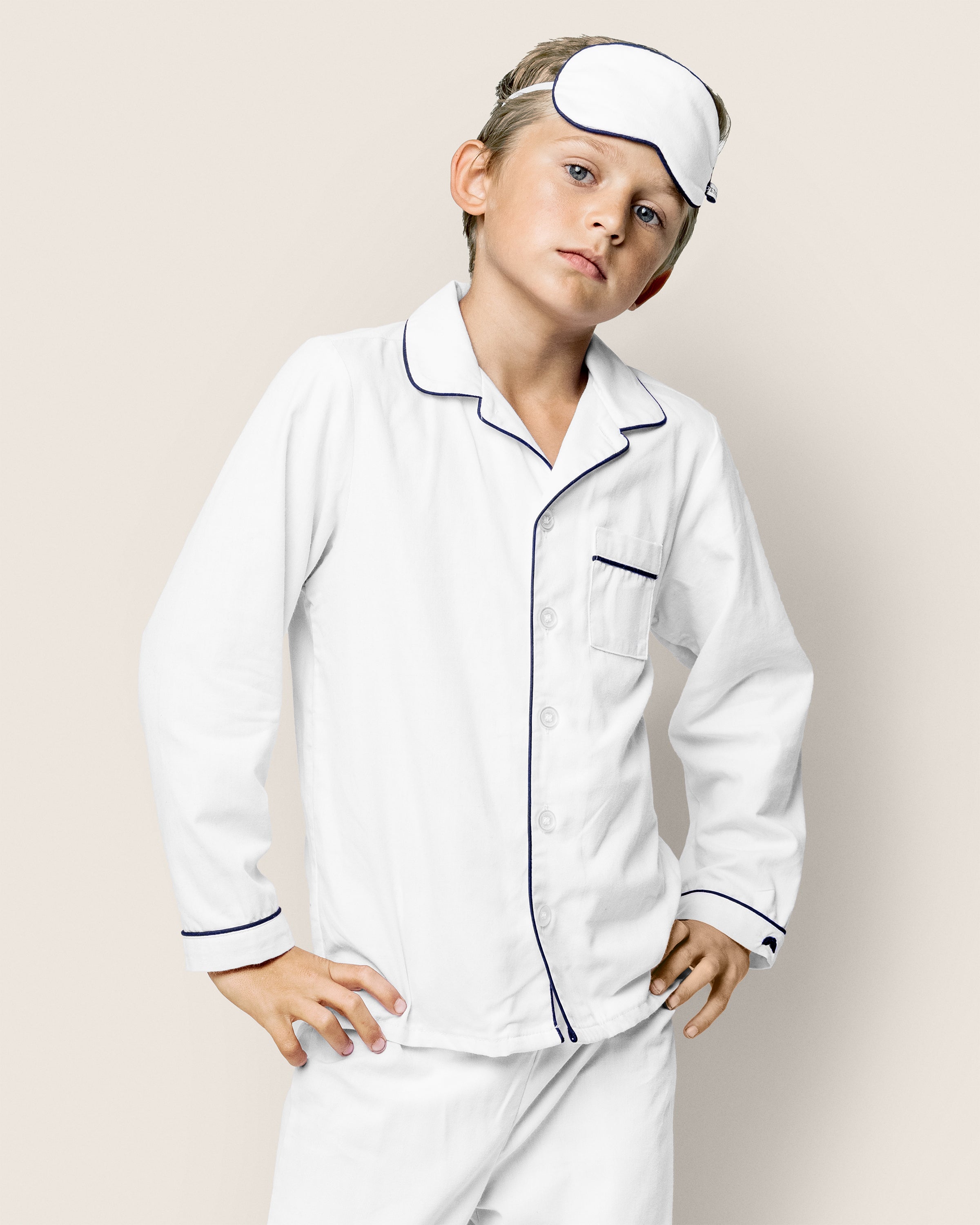 A young boy wearing Petite Plumes Kids Twill Pajama Set in White with Navy Piping and an eye mask stands confidently with his hands on his hips against a plain beige background.