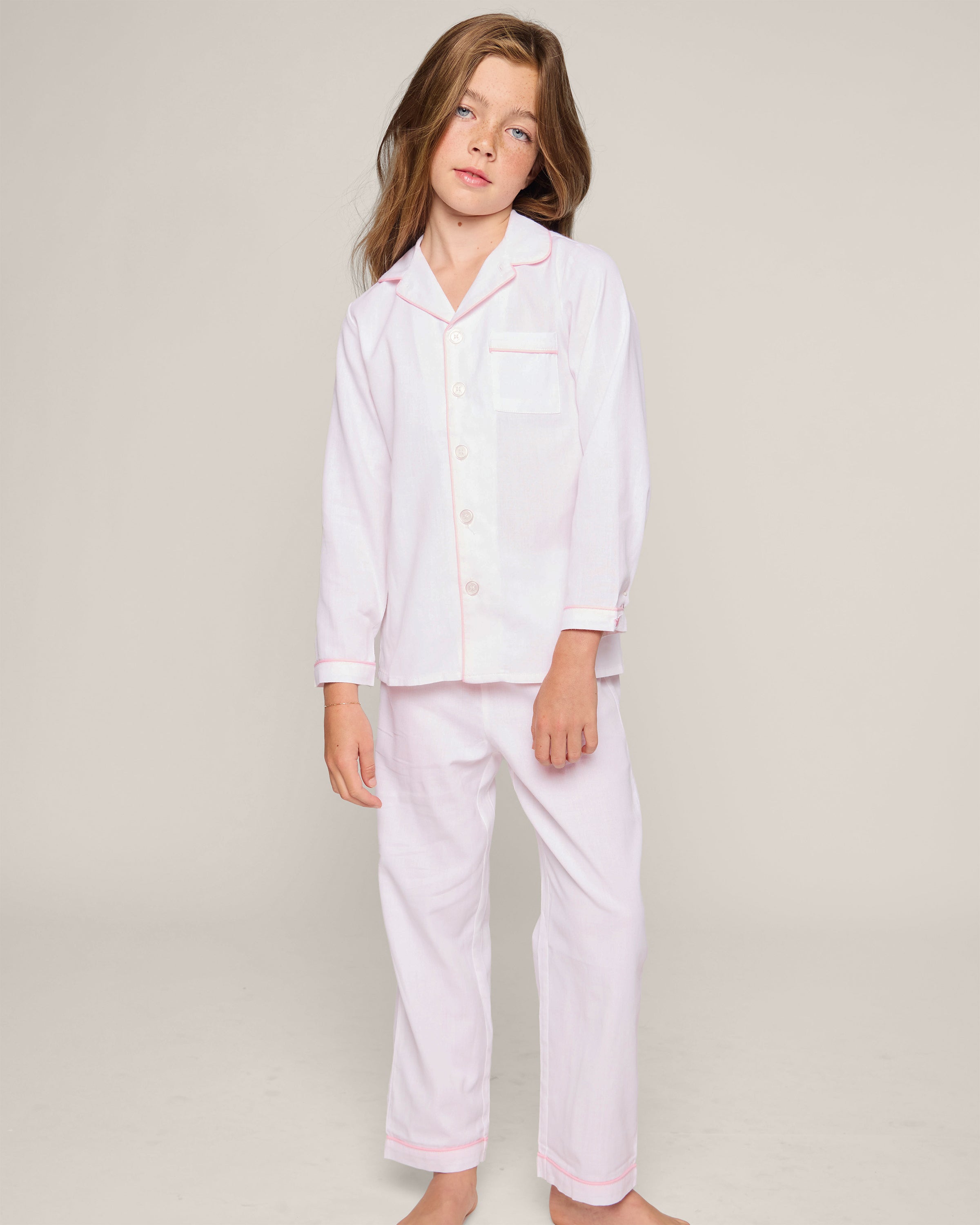 A young girl with long brown hair wears Petite Plumes Kids Twill Pajamas in white with pink piping, crafted from premium cotton. The set features a button-up shirt with a pocket and matching pants, creating a luxurious sleepwear look against a plain neutral background.