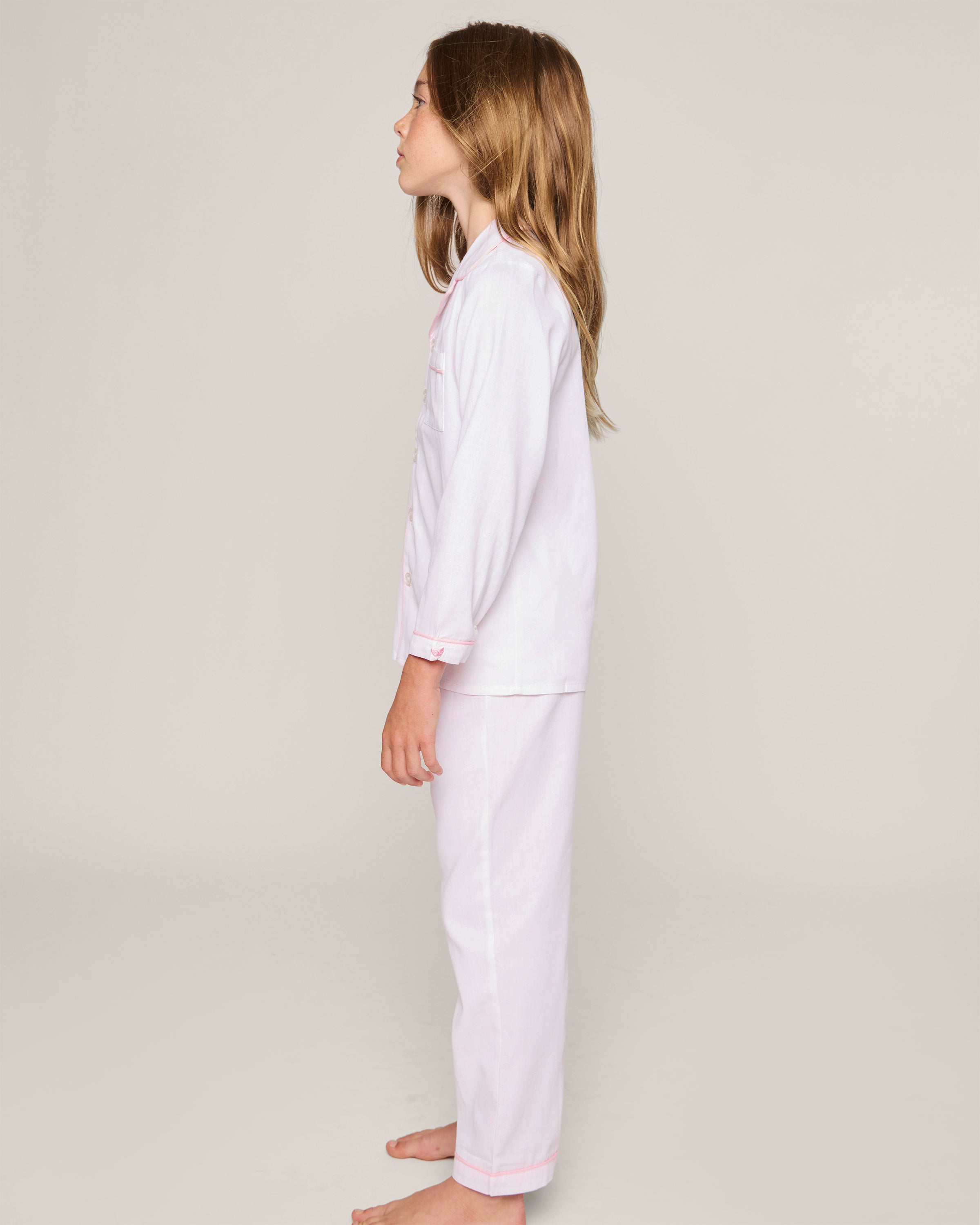 A young girl with long hair stands in profile against a plain background, wearing Petite Plumes Kids Twill Pajamas in White with Pink Piping. Crafted from high-quality cotton, she is barefoot.