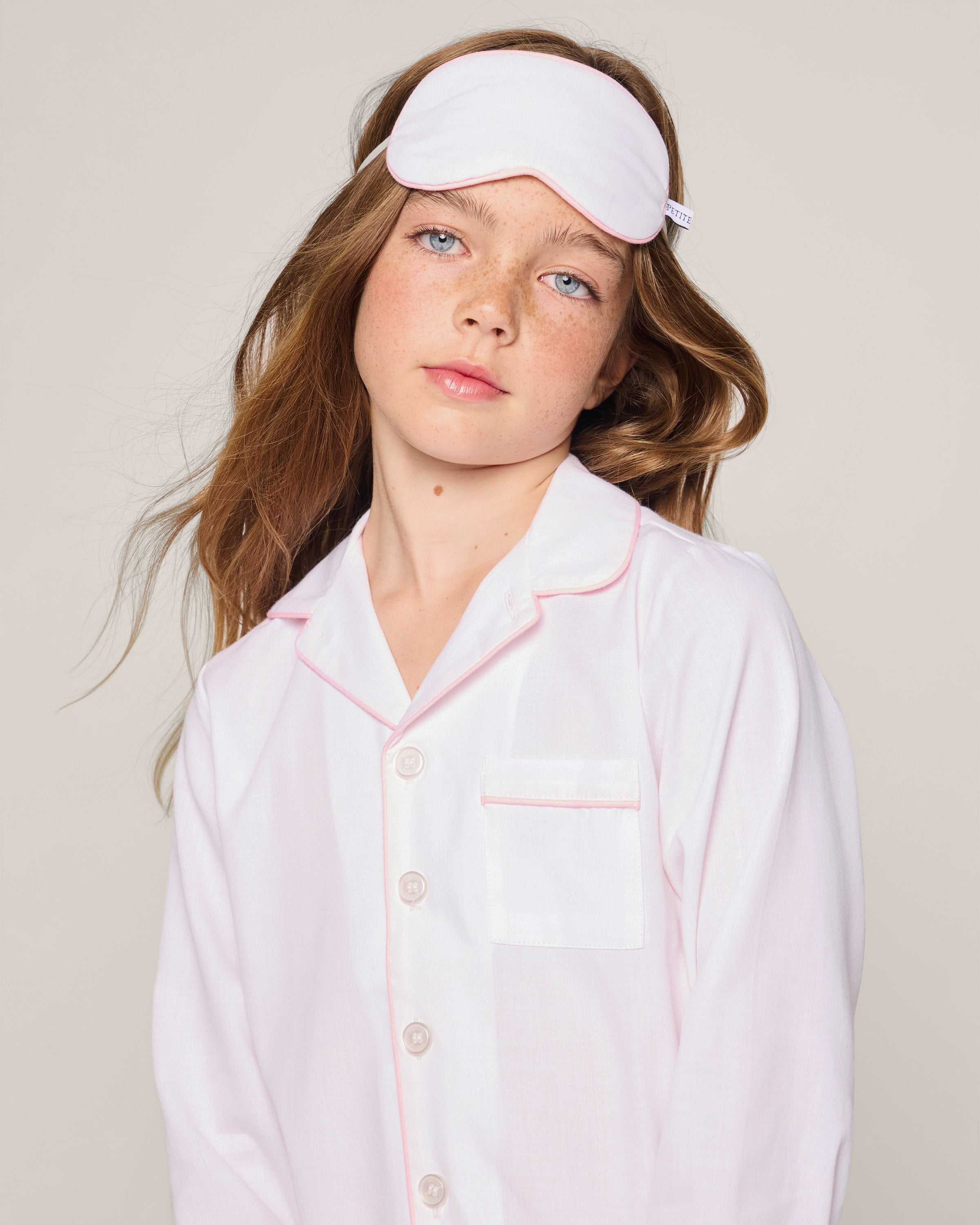 A young person with long brown hair wears a pink sleep mask and Petite Plumes Kids Twill Pajamas in white, adorned with pink piping, featuring buttons and a pocket, set against a plain background.