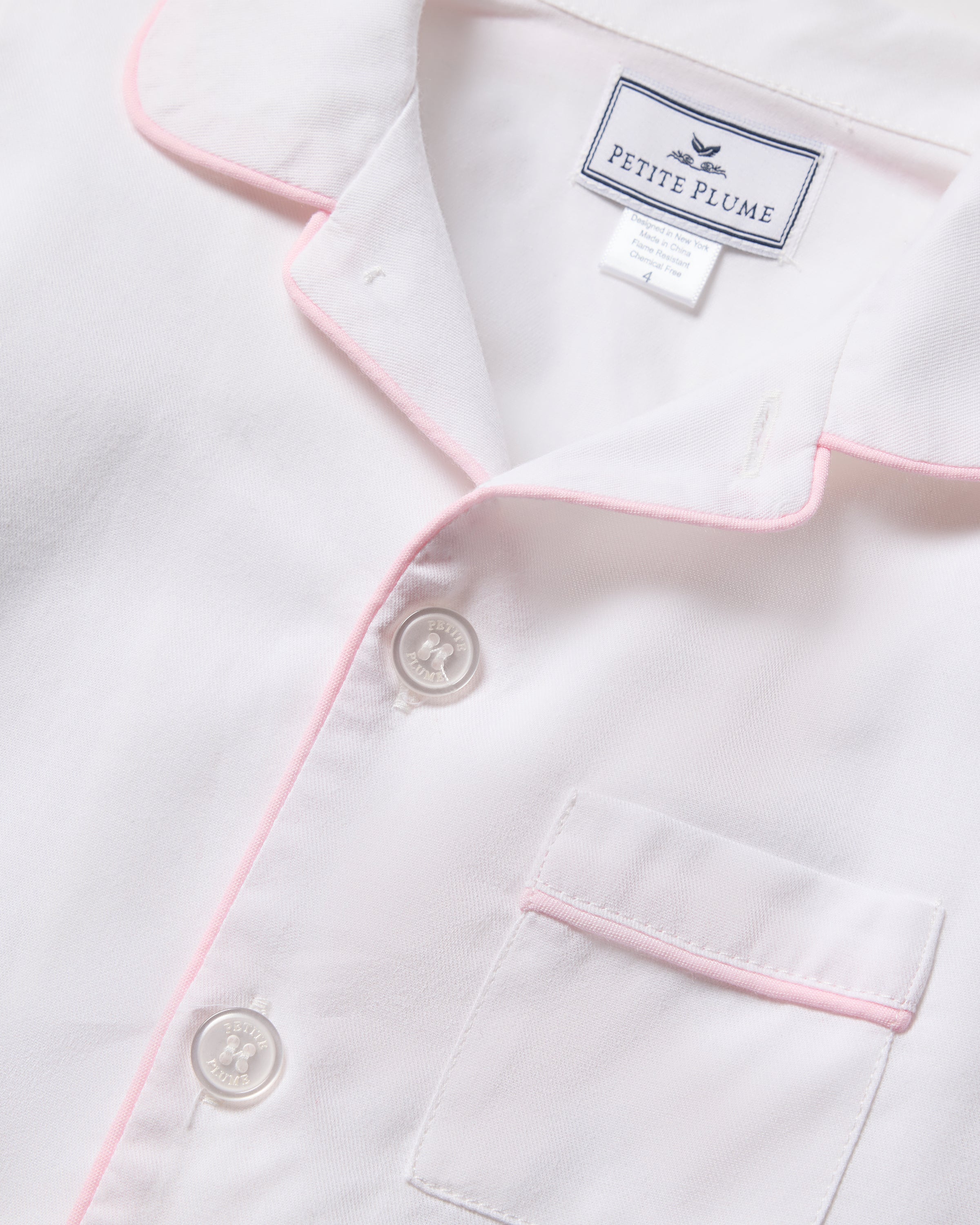 A detailed view of a luxurious Kids Twill Pajama top in white with pink piping, showcasing buttons and a small chest pocket. Petite Plume is visible on the collar label, crafted from premium cotton for comfort and elegance in this classic set.