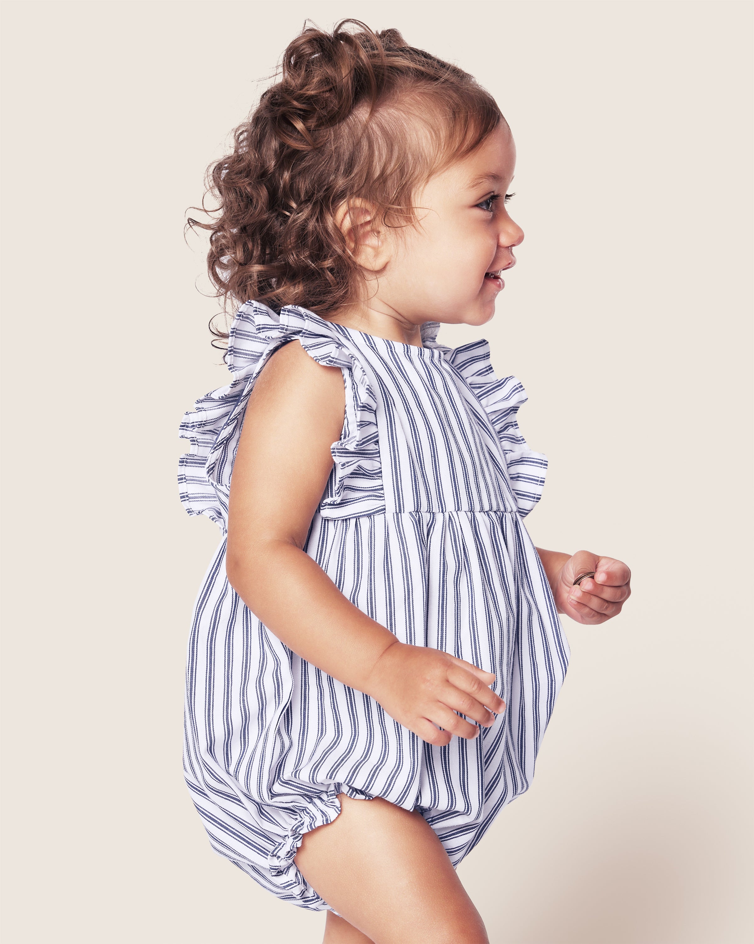 A toddler smiles in profile, wearing Petite Plumes Babys Twill Ruffled Rompers in Navy French Ticking. The yarn-dyed cotton outfit with ruffled sleeves complements the childs curly brown hair and joyful expression against a plain light background.