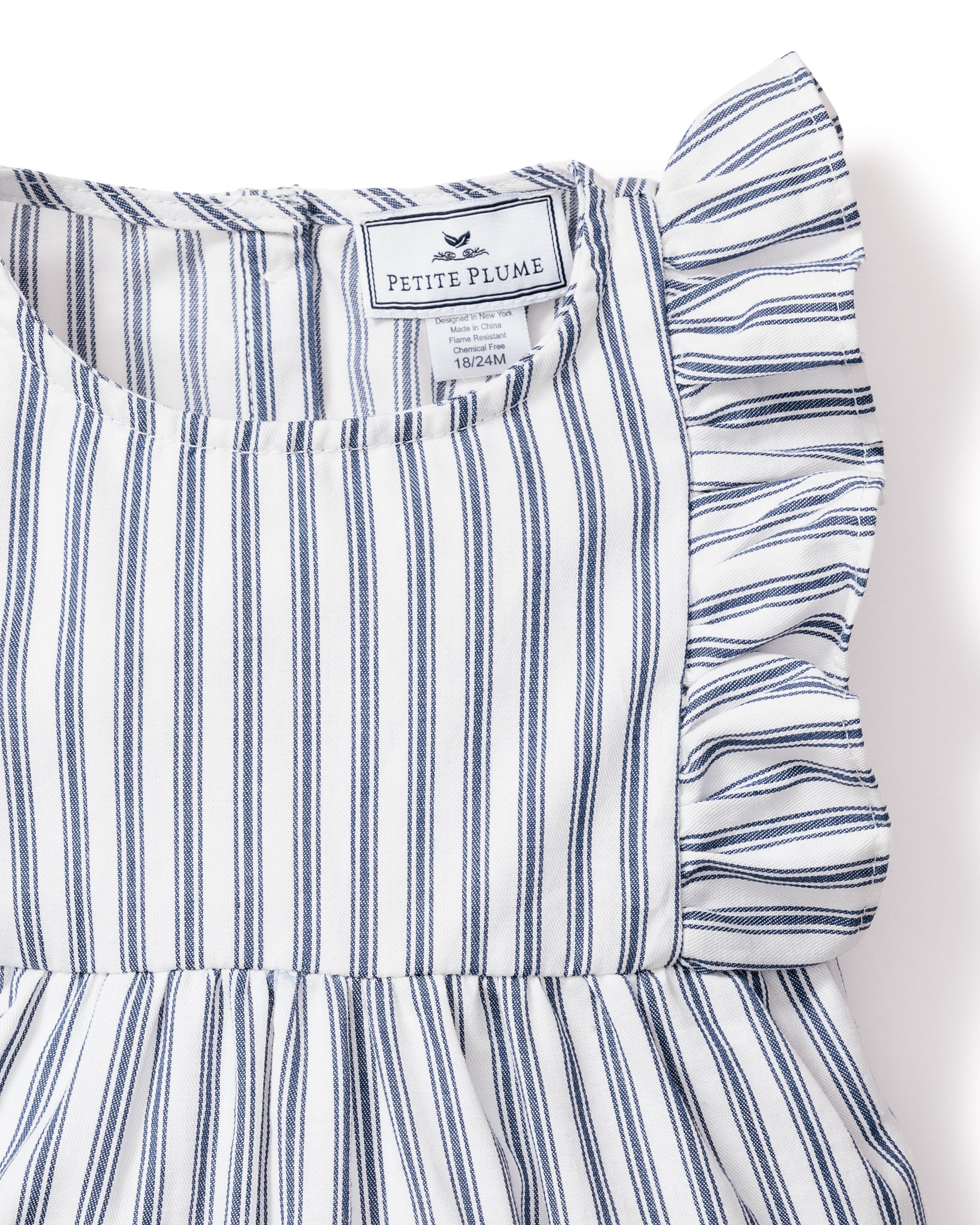 This is a Babys Twill Ruffled Romper in Navy French Ticking by Petite Plume, made from yarn-dyed cotton with blue and white stripes. It features a sleeveless design, ruffled shoulders, and a round neckline. A neck label shows Petite Plume and size 18/24M.