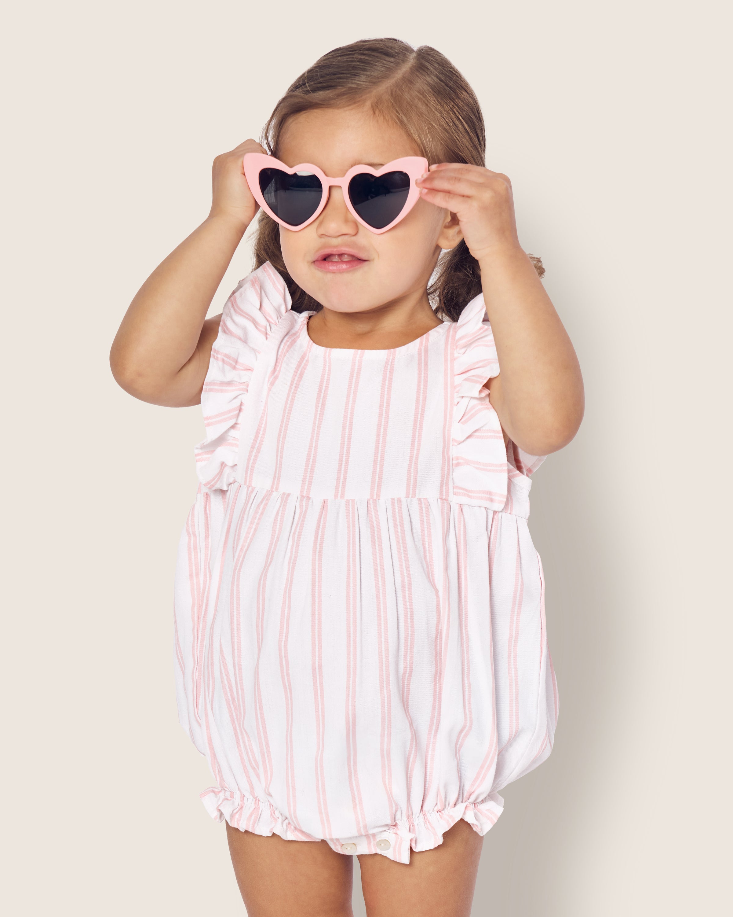 A toddler with shoulder-length brown hair, wearing heart-shaped sunglasses, adjusts them while dressed in Petite Plumes Babys Twill Stripe Ruffled Romper in Pink and White Stripe. The outfit has ruffled sleeves and elastic legs, set against a light background.