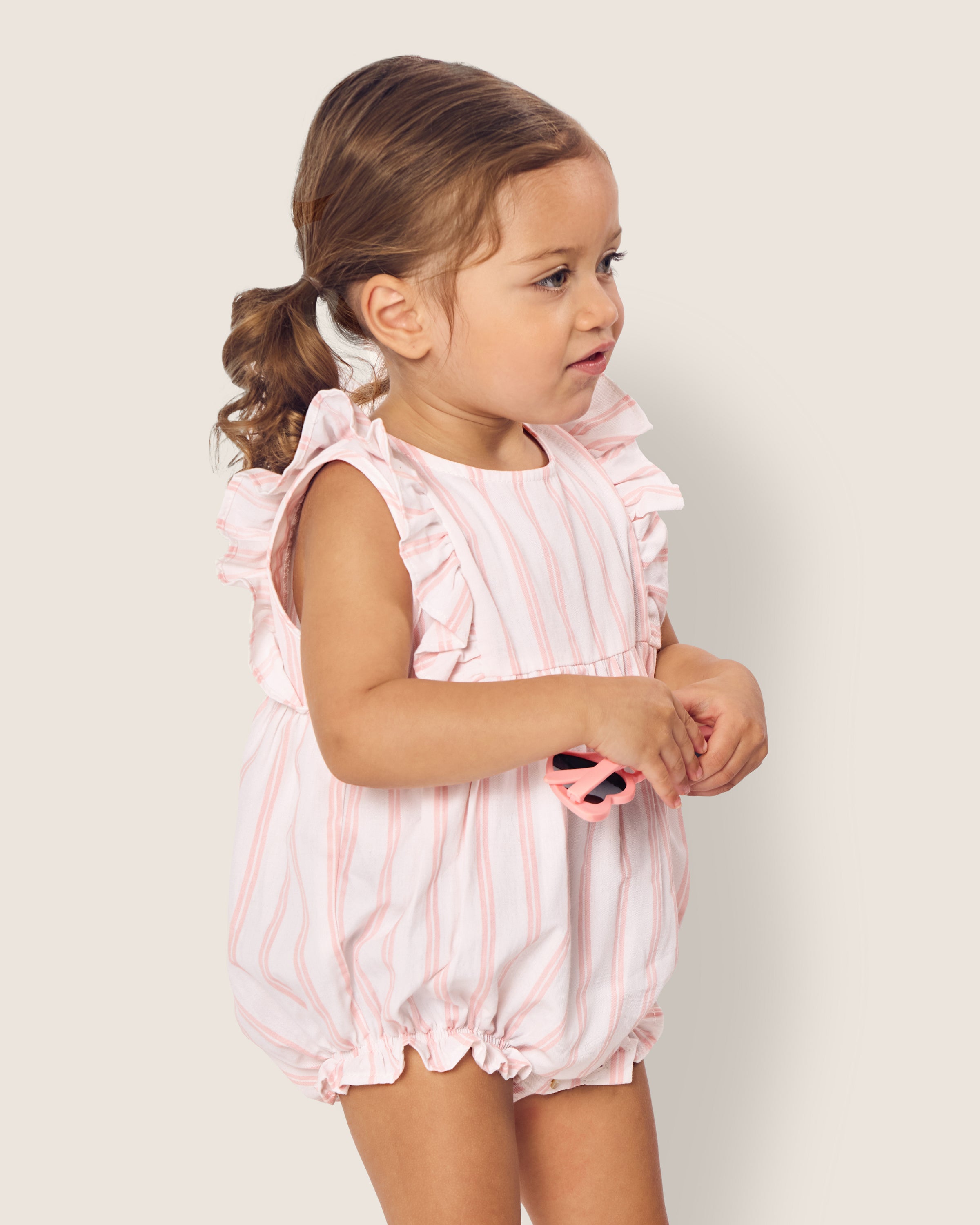 A toddler wearing Petite Plumes Babys Twill Stripe Ruffled Romper in Pink and White Stripe stands against a plain background, holding a pink pacifier. Her wavy brown hair is pulled back.