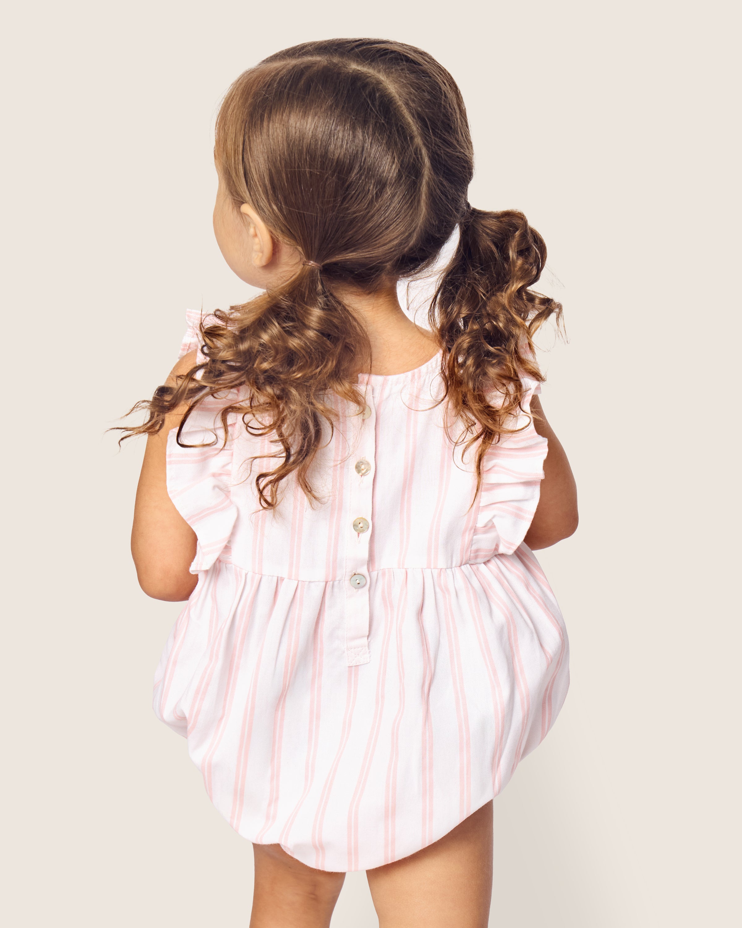 A young child with wavy brown hair is seen from behind, clad in Petite Plumes Babys Twill Stripe Ruffled Romper, featuring light pink and white stripes with ruffled sleeves. Made from yarn-dyed cotton, the romper has back buttons. The background is plain and light-colored.