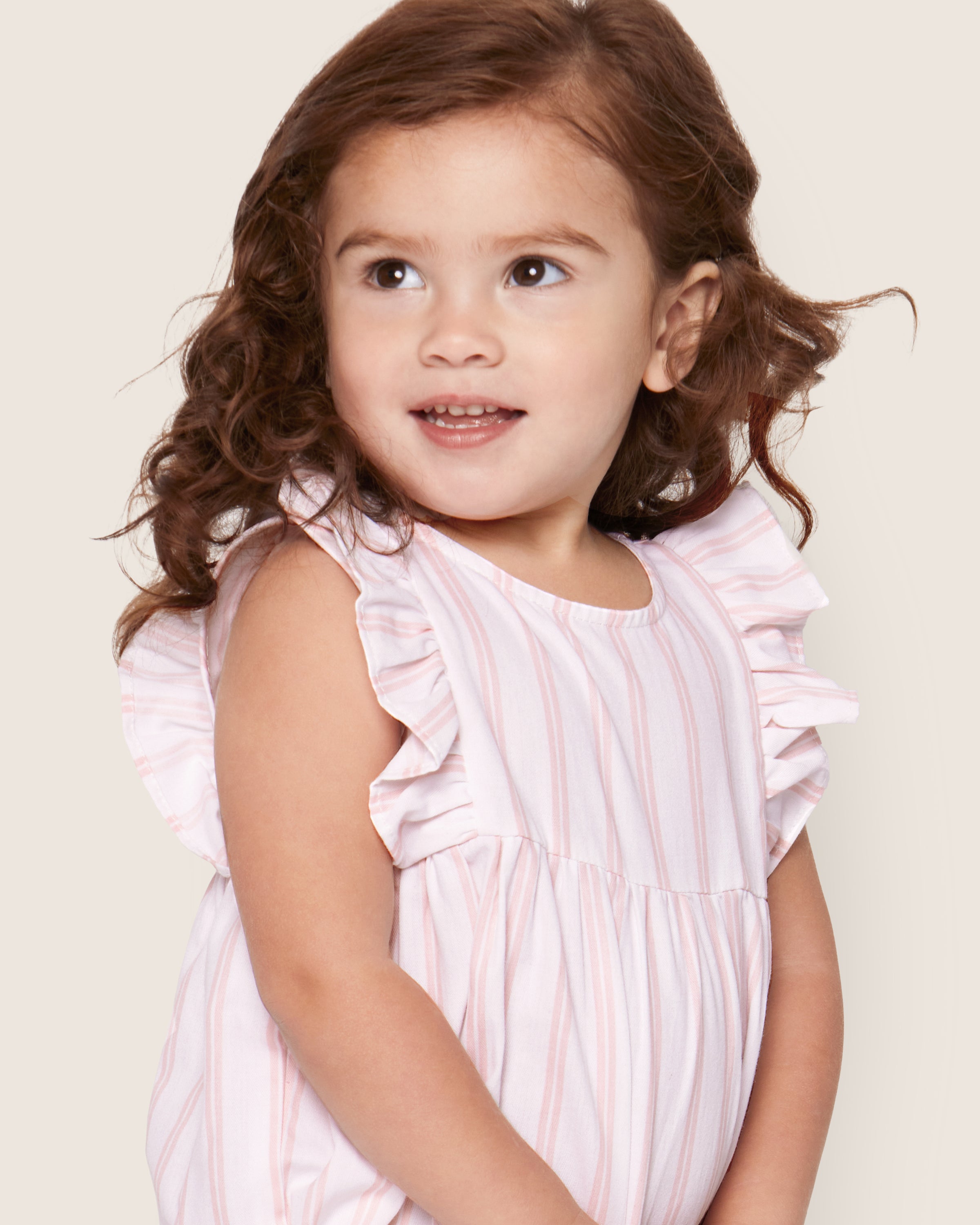 A young child with curly brown hair smiles in the Petite Plume Babys Twill Stripe Ruffled Romper, featuring pink and white stripes and ruffled sleeves, set against a soft beige background.