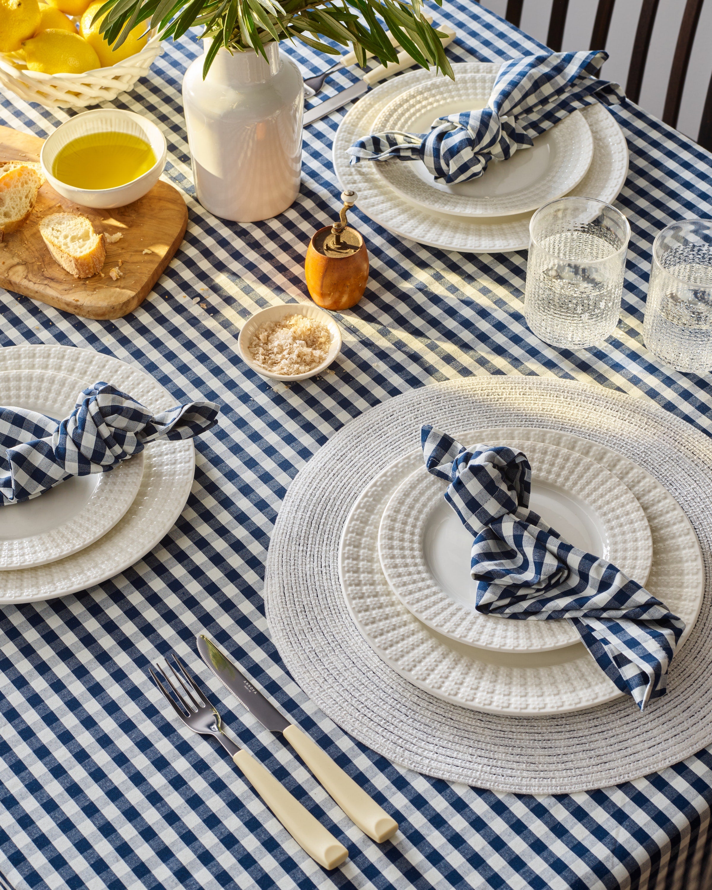 A dining table set features Signature Twill Napkins in Navy Gingham from Petite Plume, 100% cotton white plates, silverware, glasses, and a vase with plants. It includes a wooden board with bread, a small bowl of oil, and a bowl of salt for a festive tablescape.