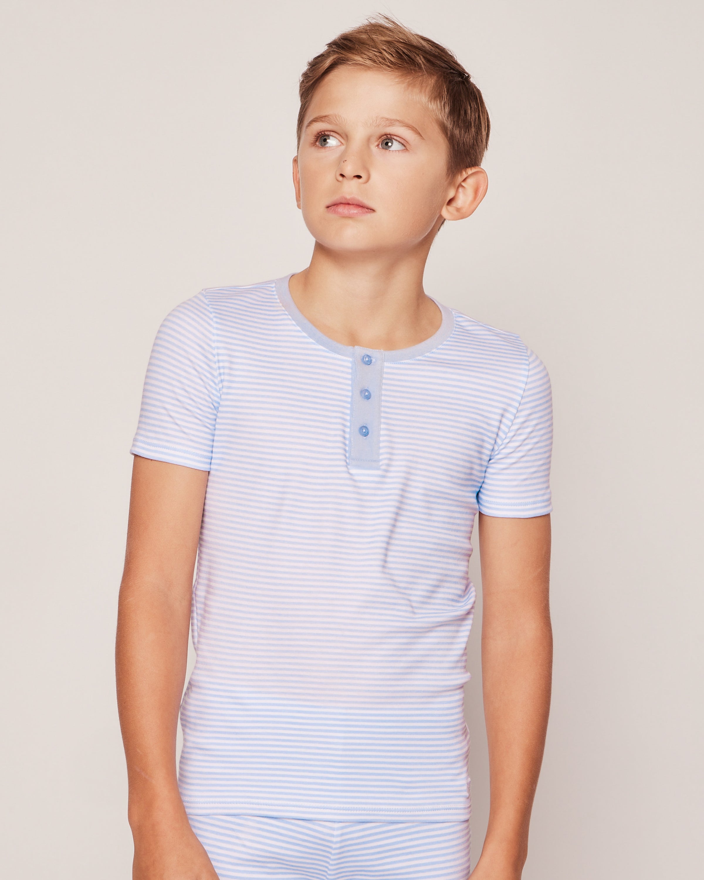 A boy in Petite Plumes Kids Pima Snug Fit Pajama Short Set, featuring light blue and white stripes with a buttoned collar, gazes to the side against a plain background.