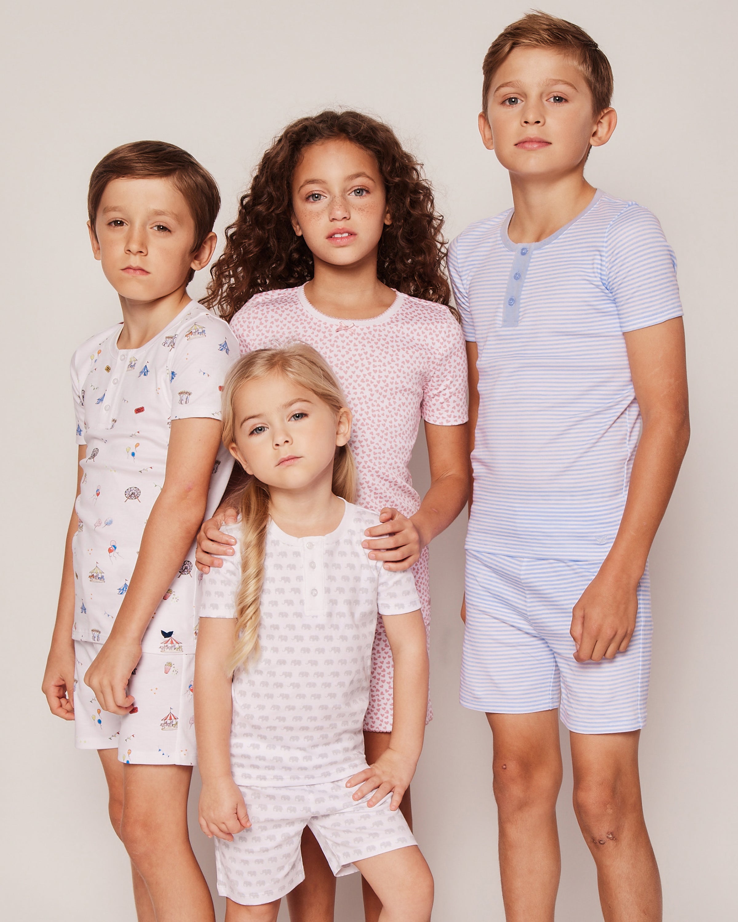 Four children pose in cozy Petite Plume pajamas, each wearing matching short-sleeved top and shorts made from soft Pima cotton. The girl in front wears a pink set, while the others showcase blue stripes and a mix of white and pastel colors against a neutral background.