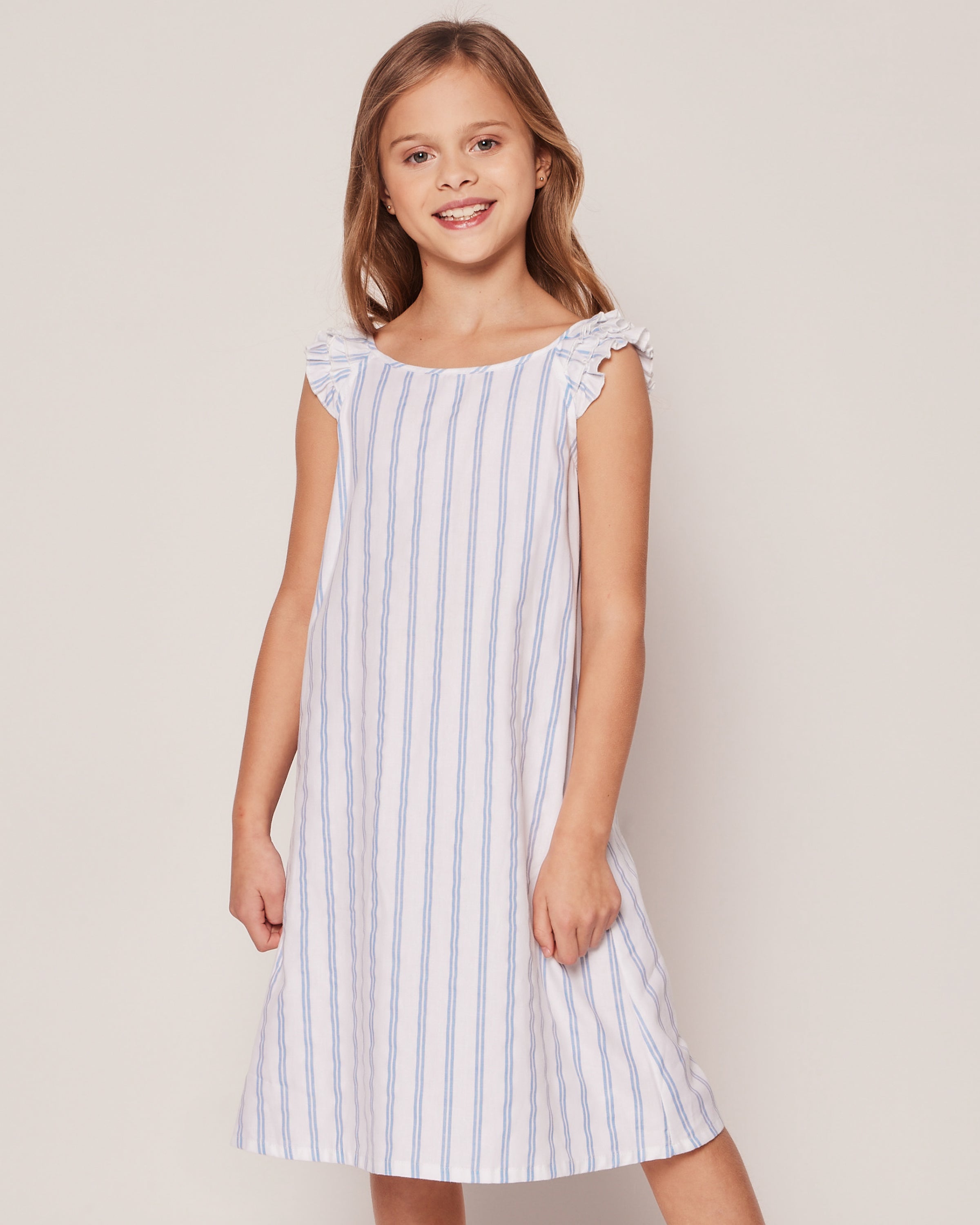 A young girl with long brown hair wears the Petite Plume Girls Twill Amelie Nightgown in a periwinkle stripe, featuring ruffled shoulders, and stands against a plain background.