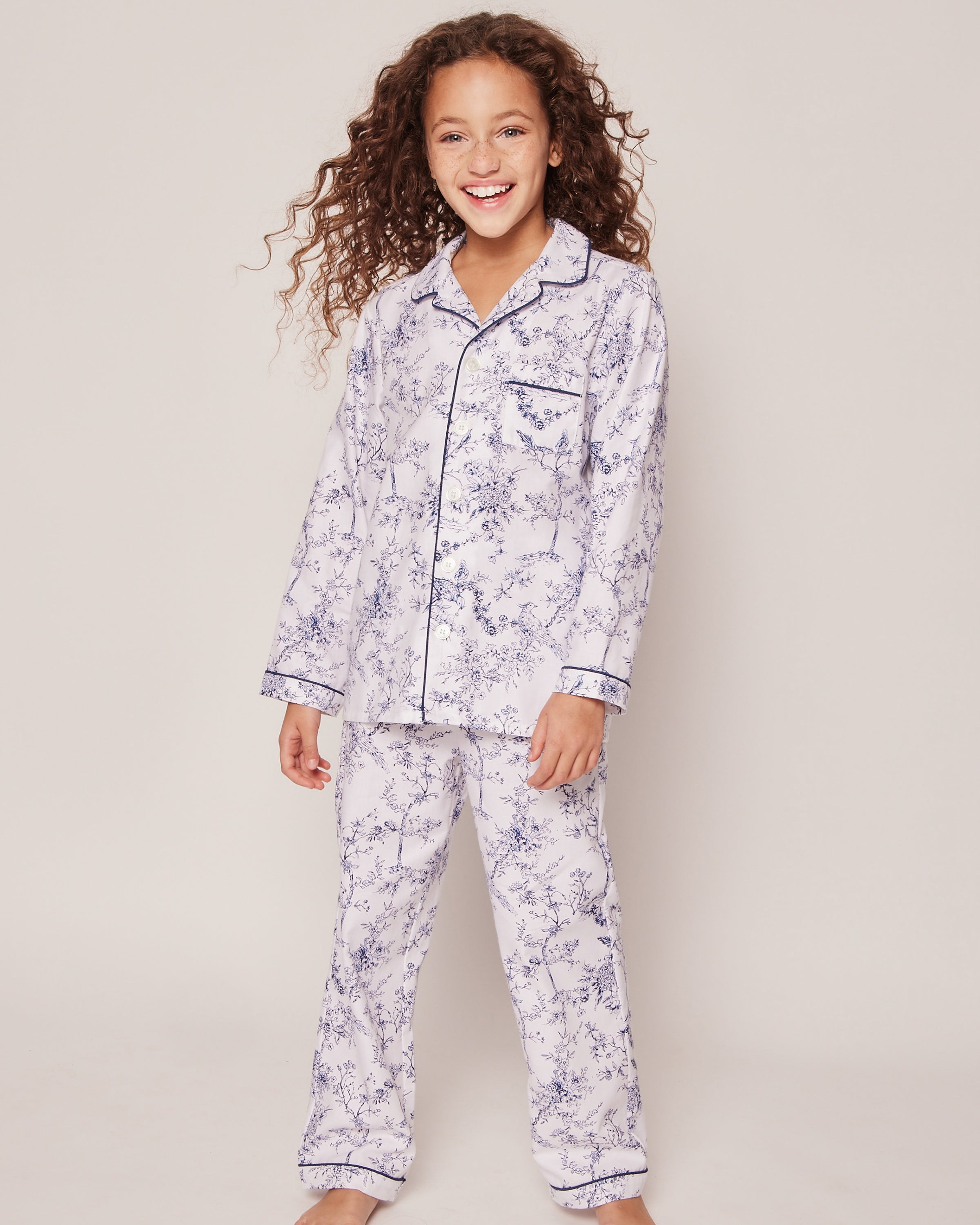 A girl with curly hair is wearing the Kids Twill Pajama Set in Timeless Toile by Petite Plume. The luxury sleepwear, made of flame retardant cotton, showcases a timeless botanical design in white with dark blue patterns and piping as she stands barefoot against a plain background.