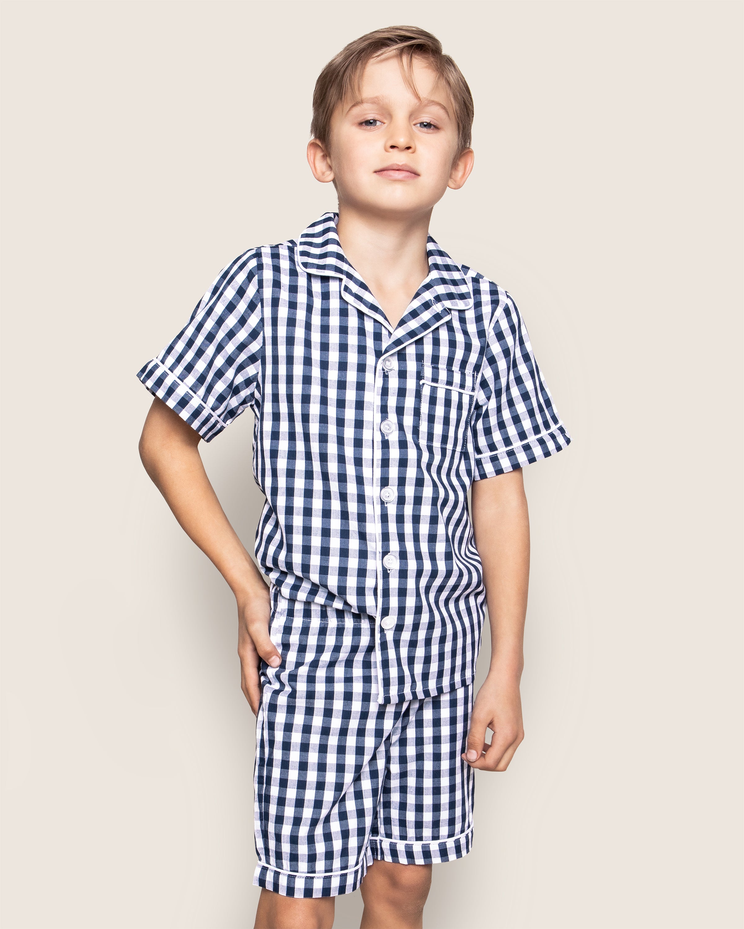 The boy confidently stands with one hand on his hip, wearing Petite Plumes Kids Pajama Short Set in Navy Gingham. The matching blue and white outfit is made from yarn-dyed cotton, ensuring both style and comfort against the light background.