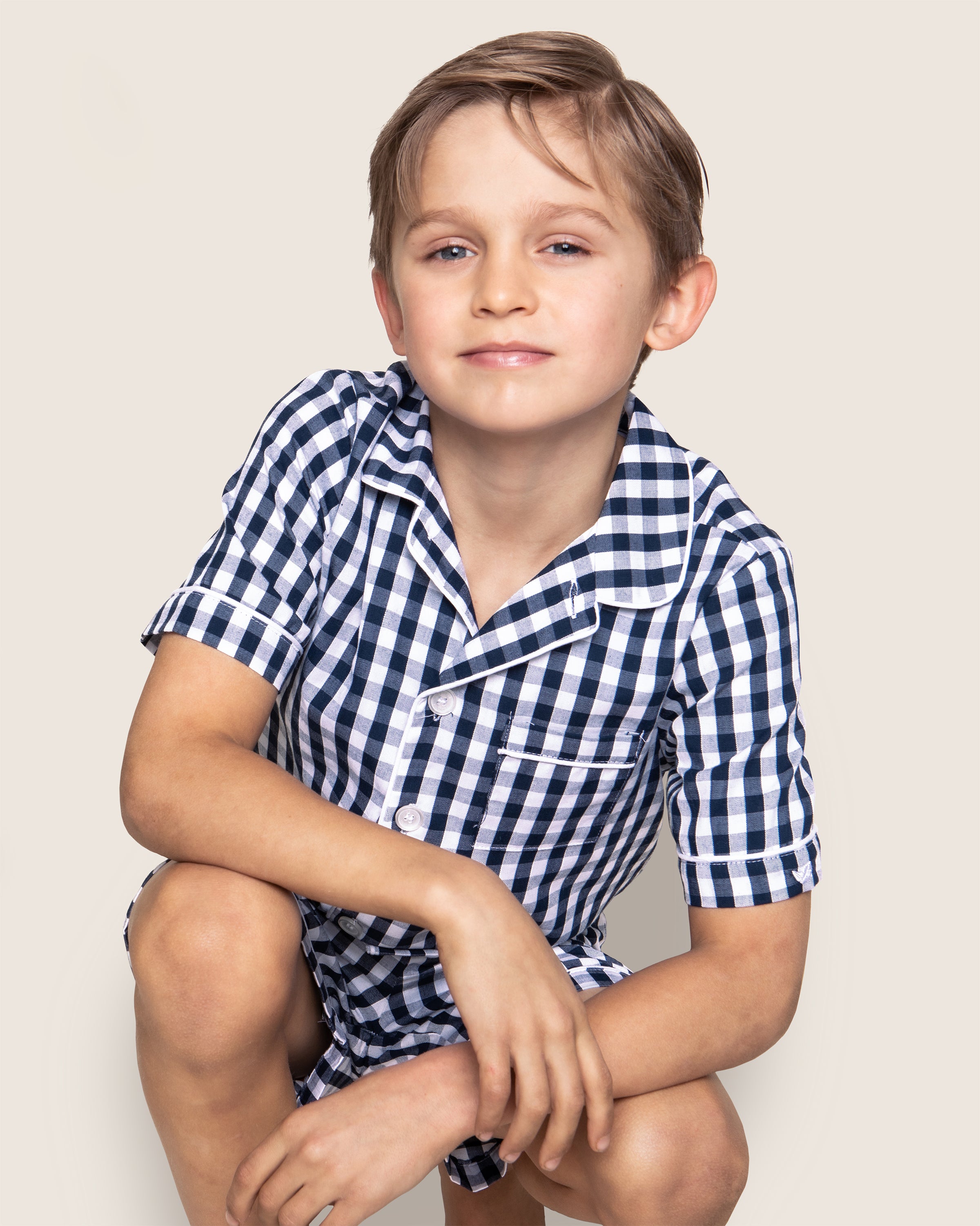 A young boy in Petite Plumes Kids Pajama Short Set in Navy Gingham crouches with one arm on his knee, highlighting the classic blue and white checkered pattern against a plain, light backdrop.