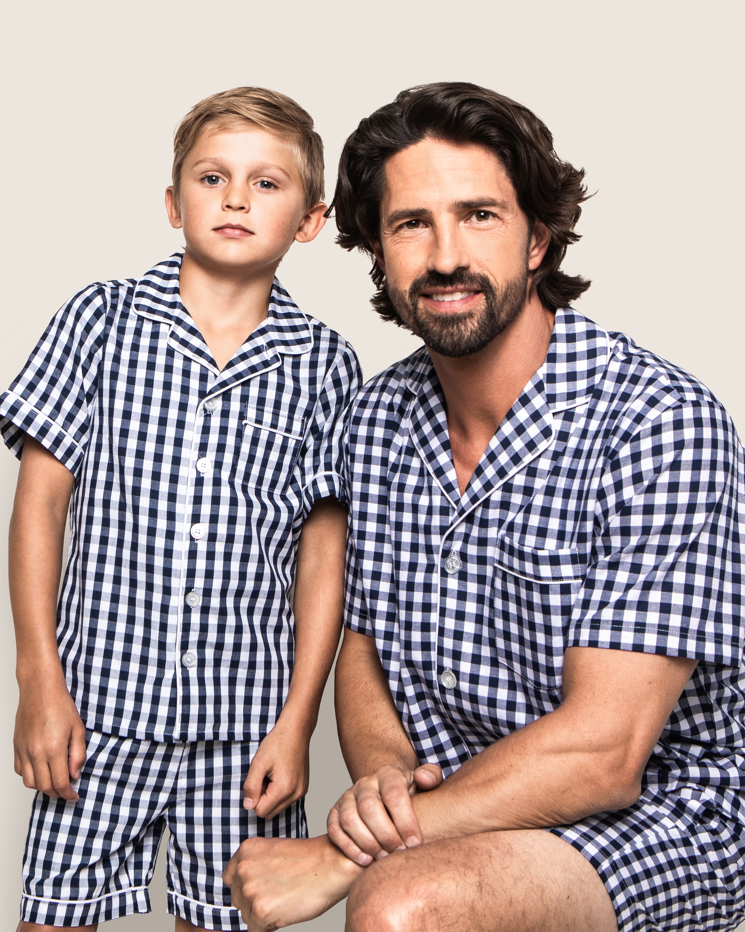 A man and boy in Petite Plumes gingham sleepwear pose against a plain background. The seated, smiling man wears cozy yarn-dyed cotton pajamas, while the boy stands with a neutral expression wearing the Kids Pajama Short Set in Navy Gingham. Both sport short hair, enhancing their charming matching look.