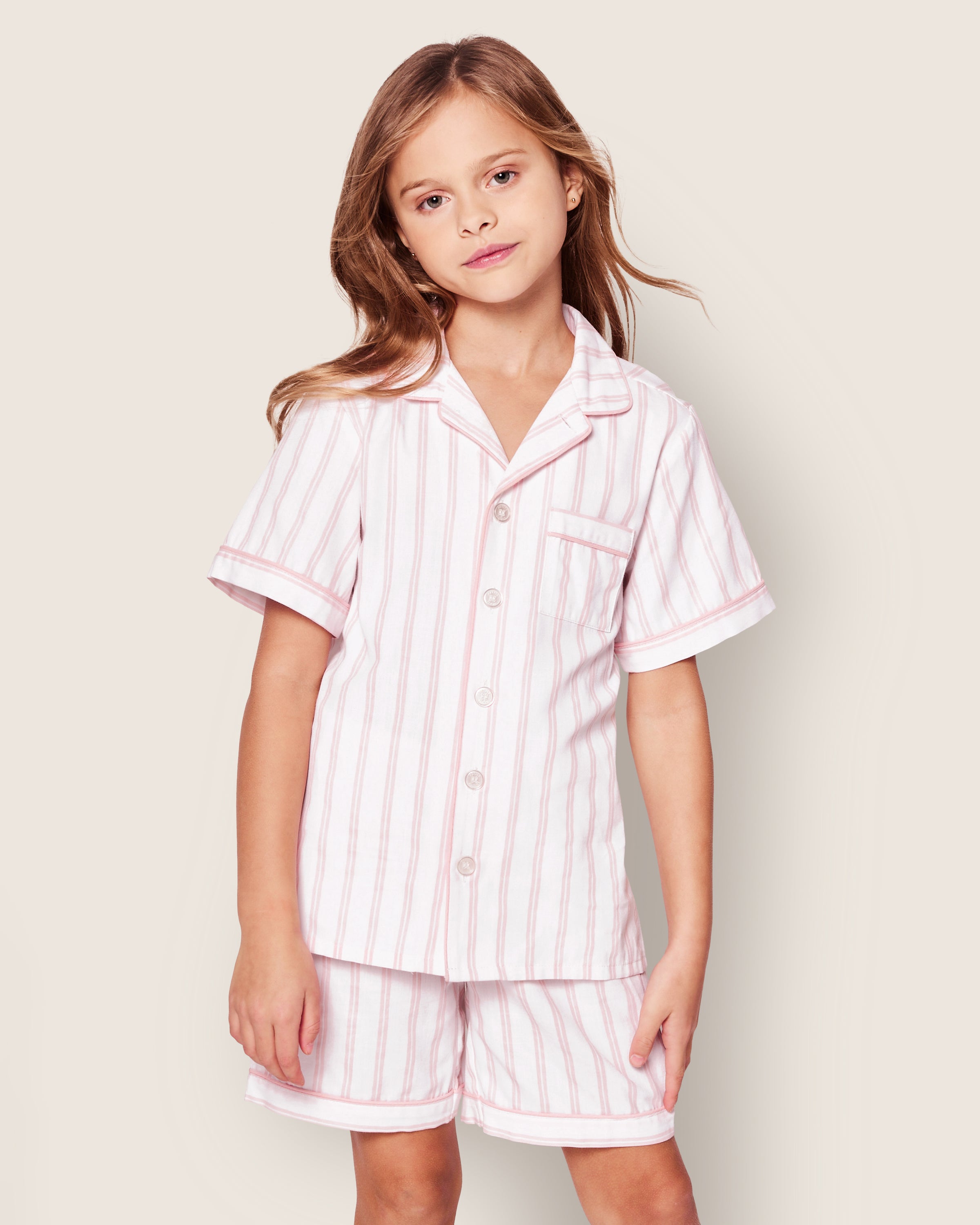 A child in the Petite Plume Kids Twill Pajama Short Set, featuring pink and white stripes, stands against a plain beige background with a relaxed expression.