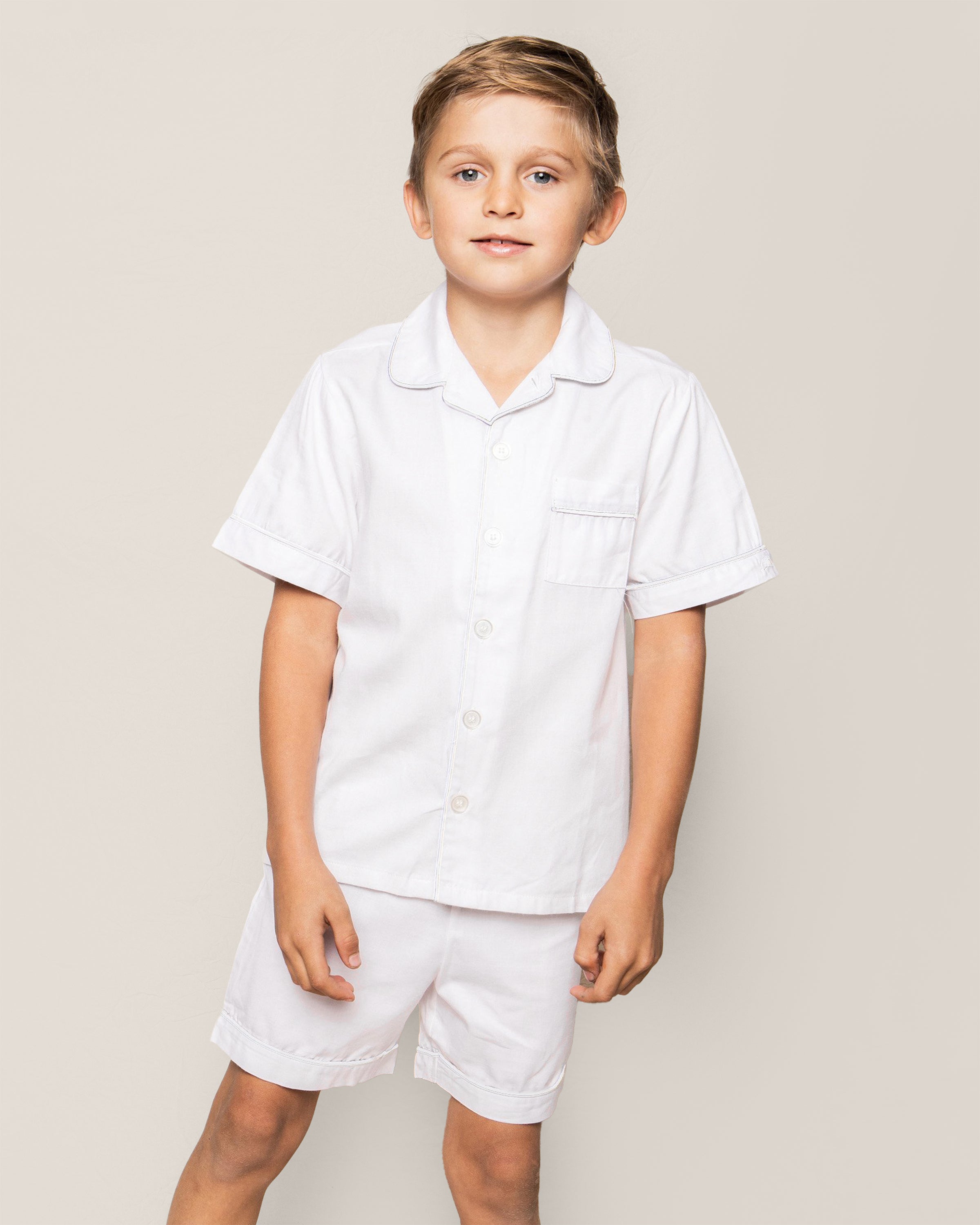 A young boy with short, light brown hair smiles at the camera against a plain background, wearing the Kids Twill Pajama Short Set in White by Petite Plume.