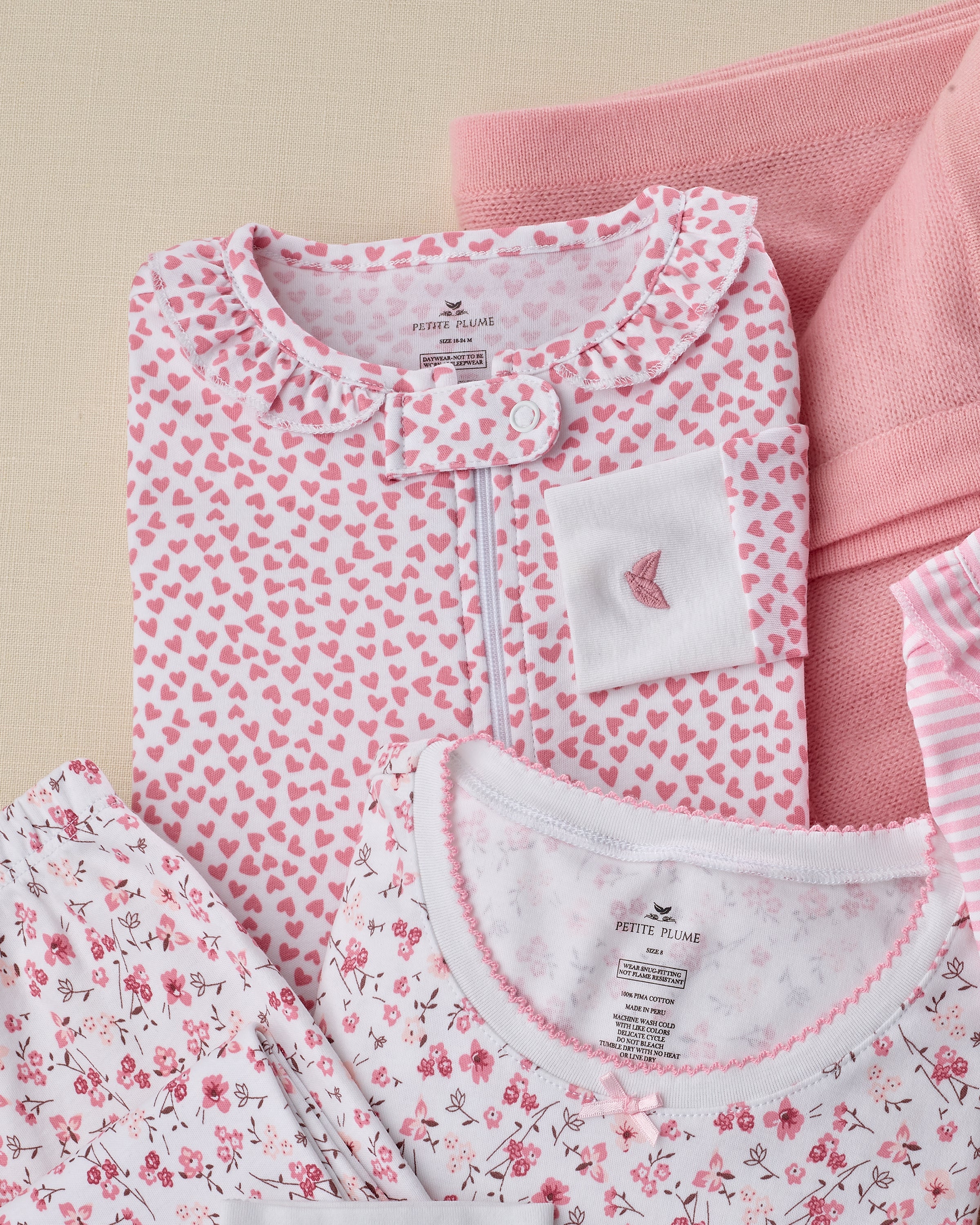 A neatly arranged stack of childrens clothing showcases Petite Plumes Baby Pima Ruffle Collar Romper in Sweethearts with pink and white heart and floral patterns. The set includes a top with a pocket, a striped piece, on a light surface with a soft pink blanket.