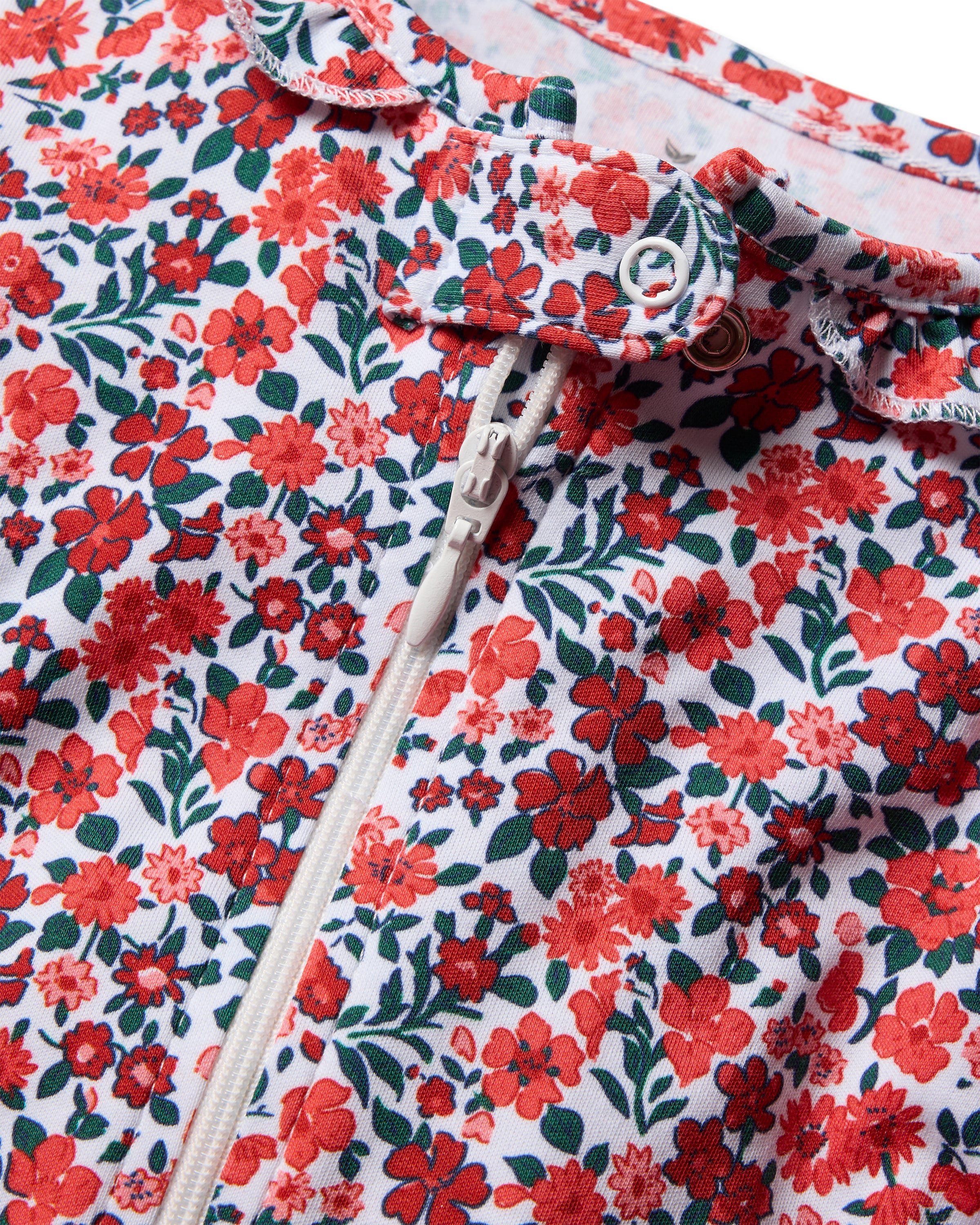 Close-up of Petite Plumes Babys Pima Ruffle Collar Romper in Fleurs des Bois. This elegant sleepwear, crafted from soft Pima cotton, features a floral pattern with red flowers and green leaves on white, a zipper, a button, and charming ruffled edges.