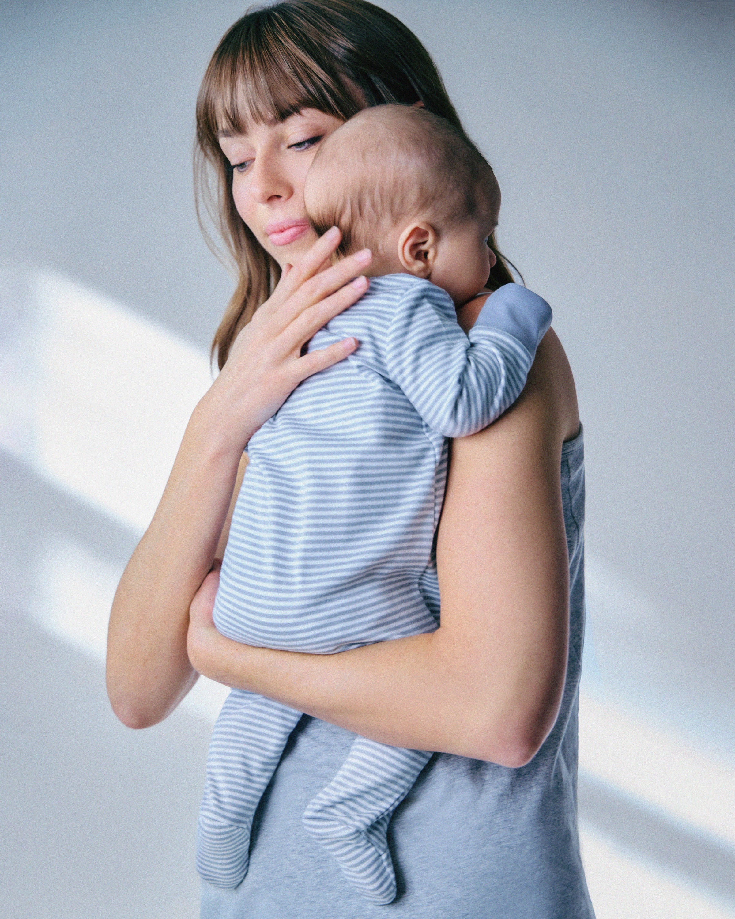 A woman with long hair gently holds a baby on her shoulder. The baby is wearing a snug-fit romper made of soft Pima cotton in grey stripes by Petite Plume. The softly lit background enhances the peaceful and intimate atmosphere.