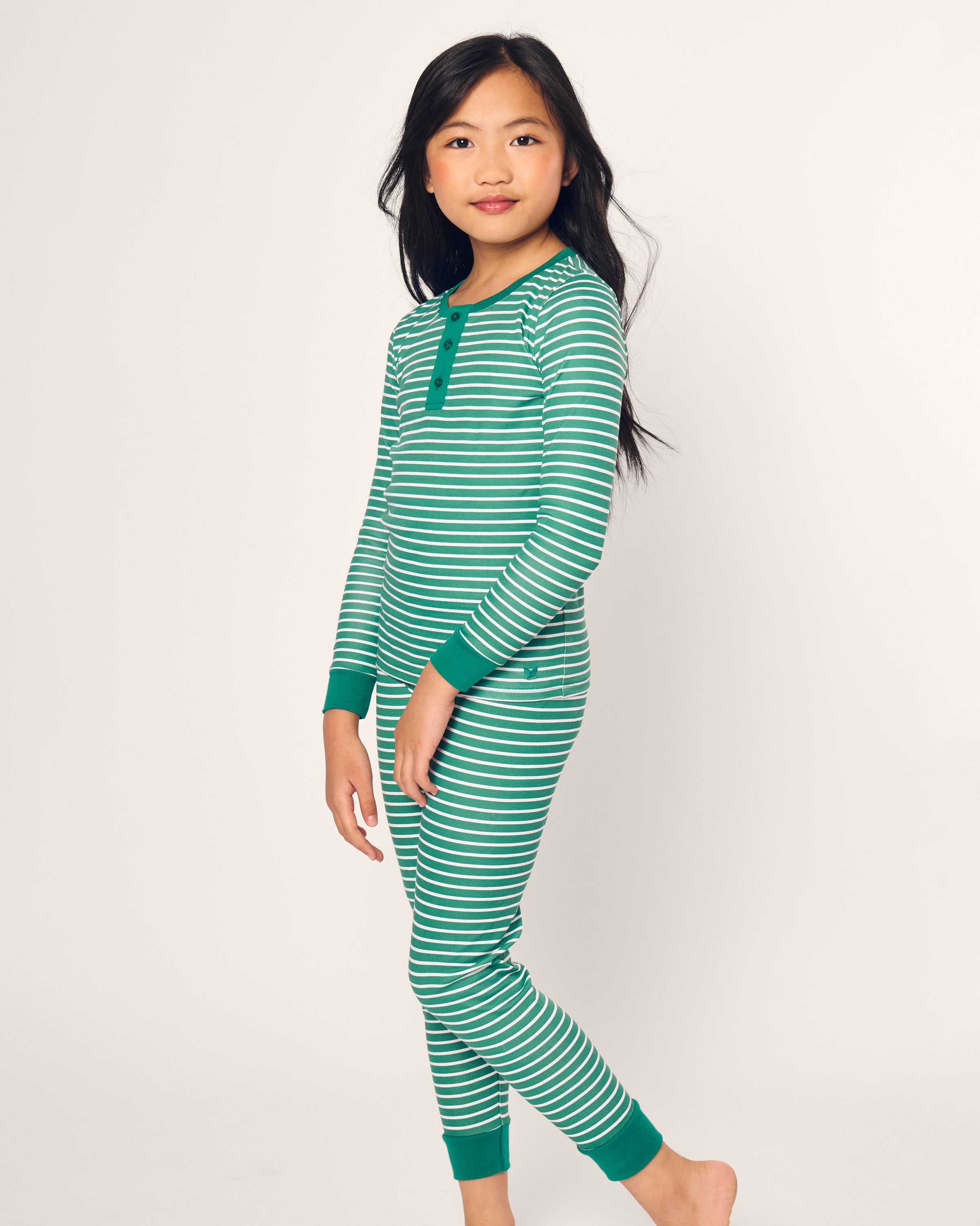 A child smiles in Petite Plumes Kids Pima Snug Fit Pajama Set in Green Stripe, featuring a green and white long sleeve top and pants with green cuffs. The pose is relaxed against a plain background, dark hair falling over the shoulders.