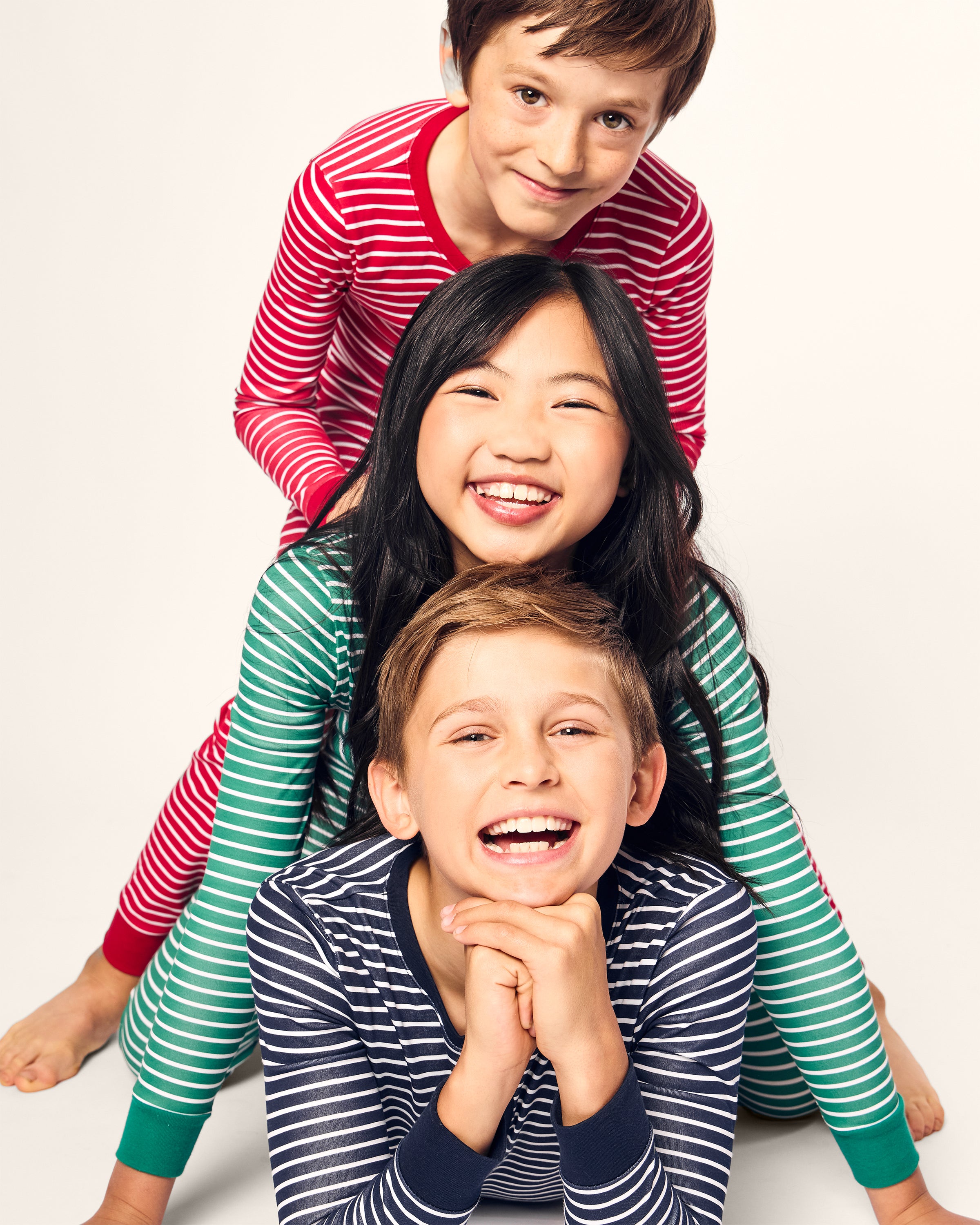 Three children in Petite Plumes cozy Pima snug fit pajama sets, smiling at the camera, are playfully stacked on each other. The top wears red stripes, the middle in green (Kids Pima Snug Fit Pajama Set in Green Stripe), and the bottom wears blue, all popping against a plain white background.