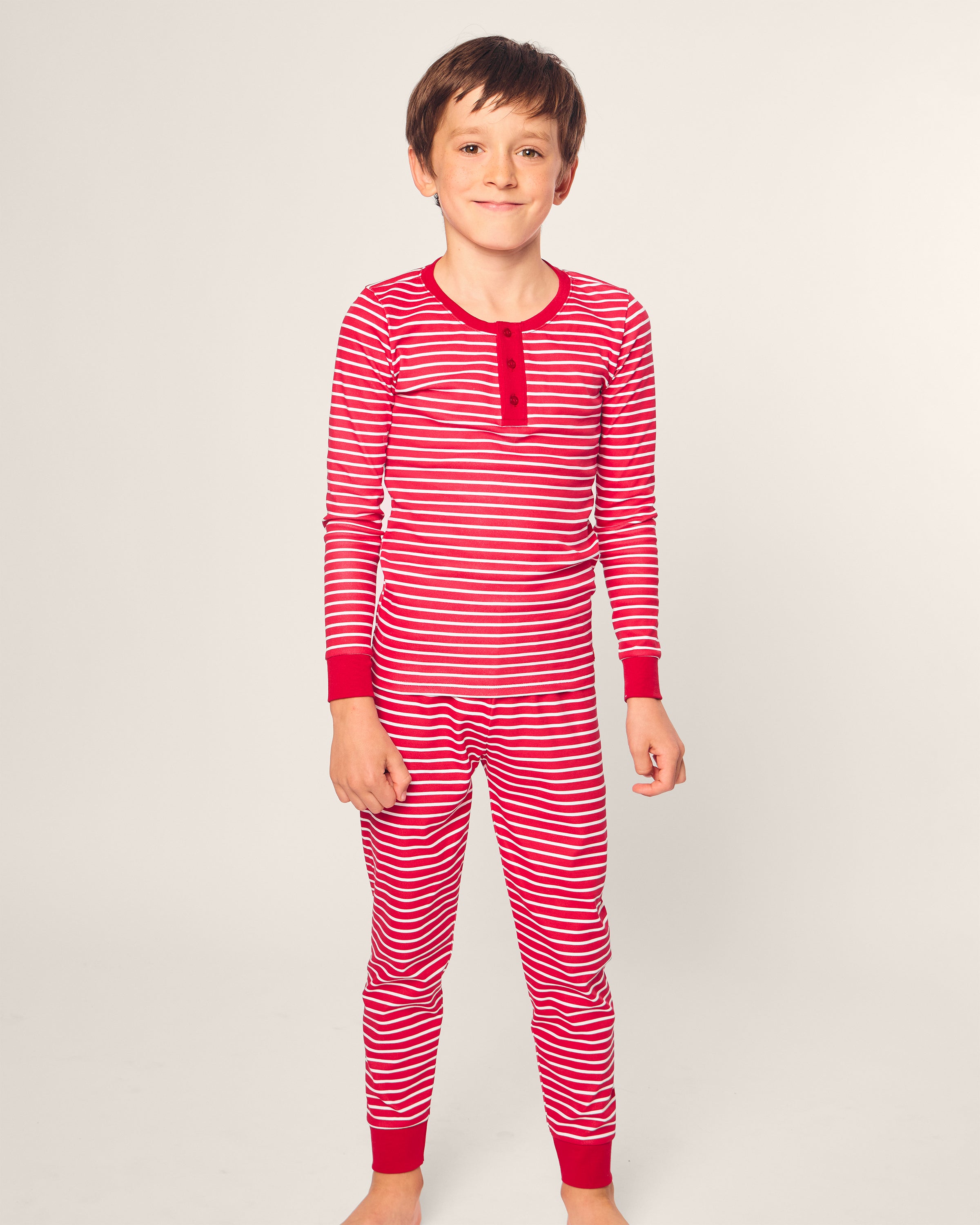 A child wearing the Kids Pima Snug Fit Pajama Set in Red Stripe by Petite Plume stands against a plain background, smiling.