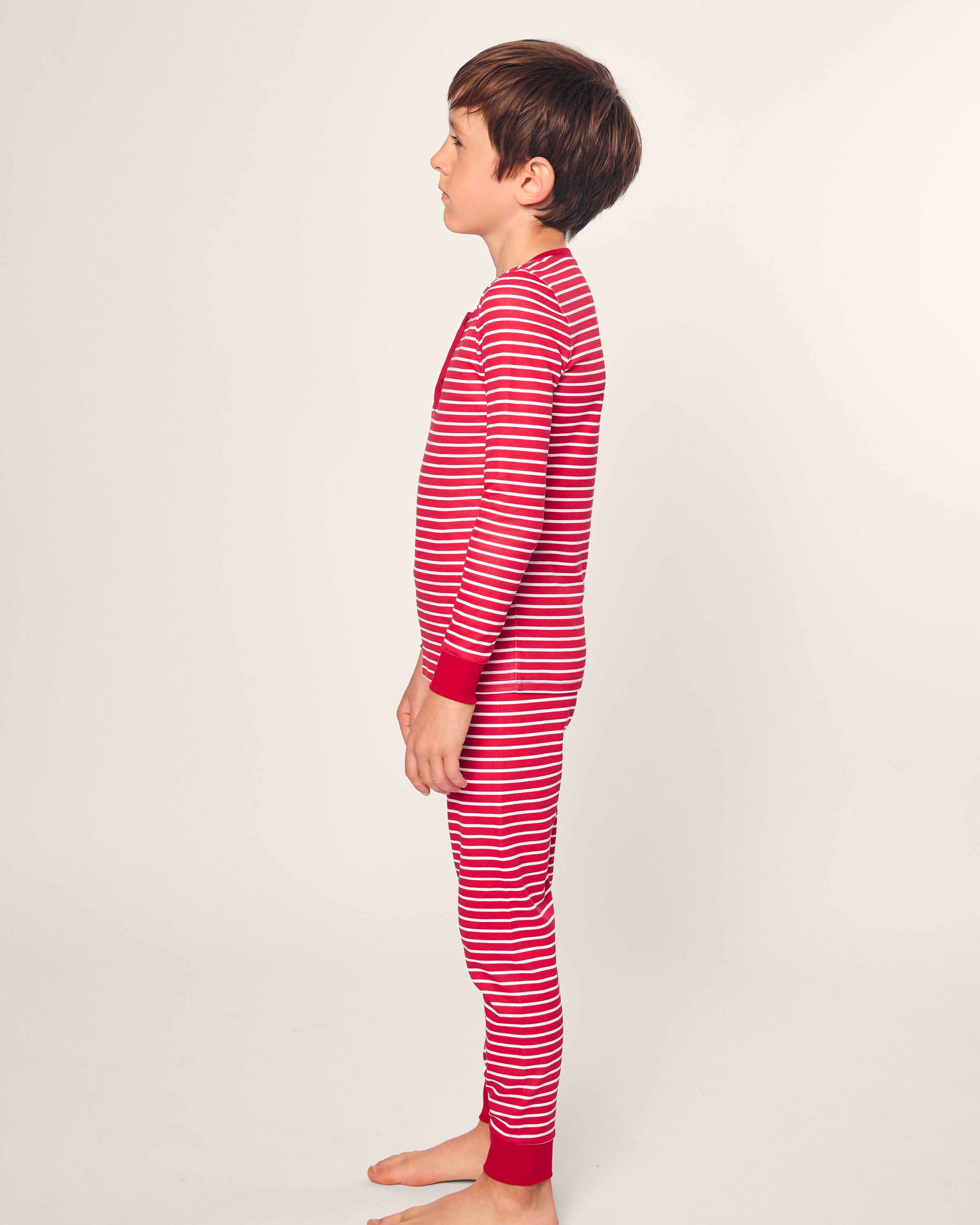 A child stands barefoot in profile against a plain background, wearing Petite Plumes Kids Pima Snug Fit Pajama Set in Red Stripe. The sleepwear features a long-sleeve top and pant set with red and white stripes and red cuffs for a cozy bedtime look.