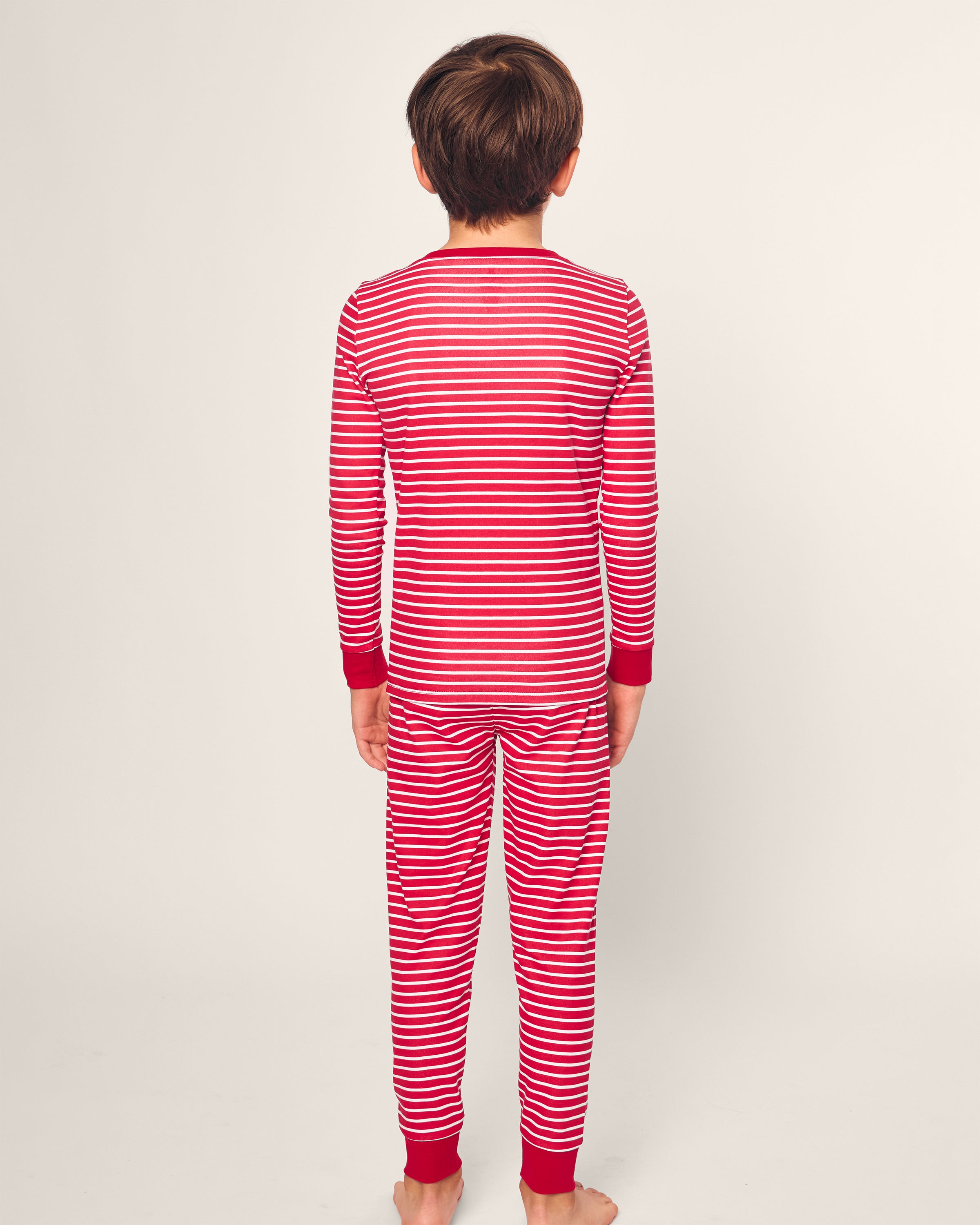 A child, seen from behind with short brown hair, stands barefoot against a plain light background wearing the Petite Plume Kids Pima Snug Fit Pajama Set in red and white stripes with cozy red cuffs.